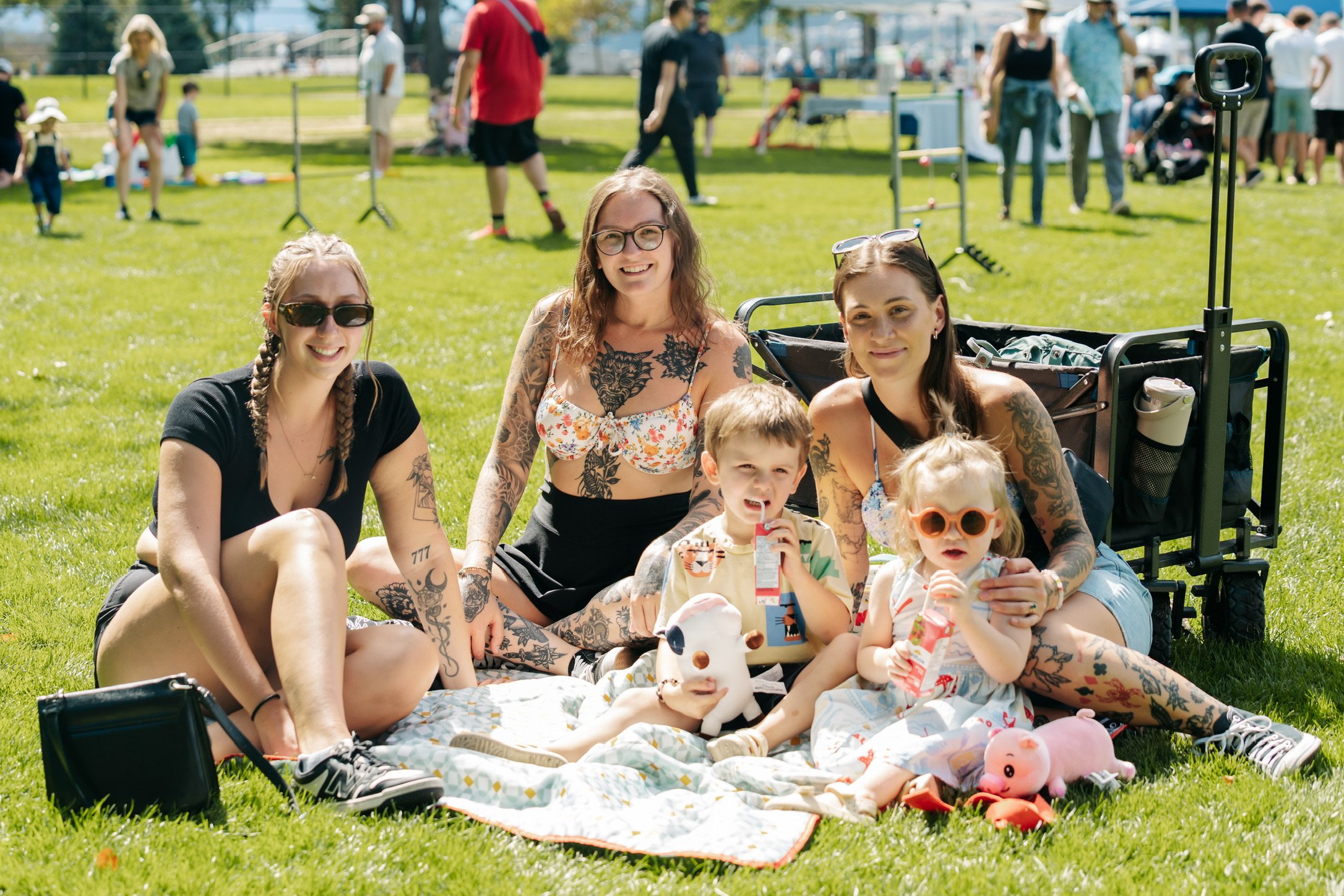 Group of five people, including children, sitting on a blanket on the grass at a park during a sunny day. They are smiling and enjoying the outdoor event, with some holding drinks and toys. The background shows other people, playground equipment, and outdoor activities.