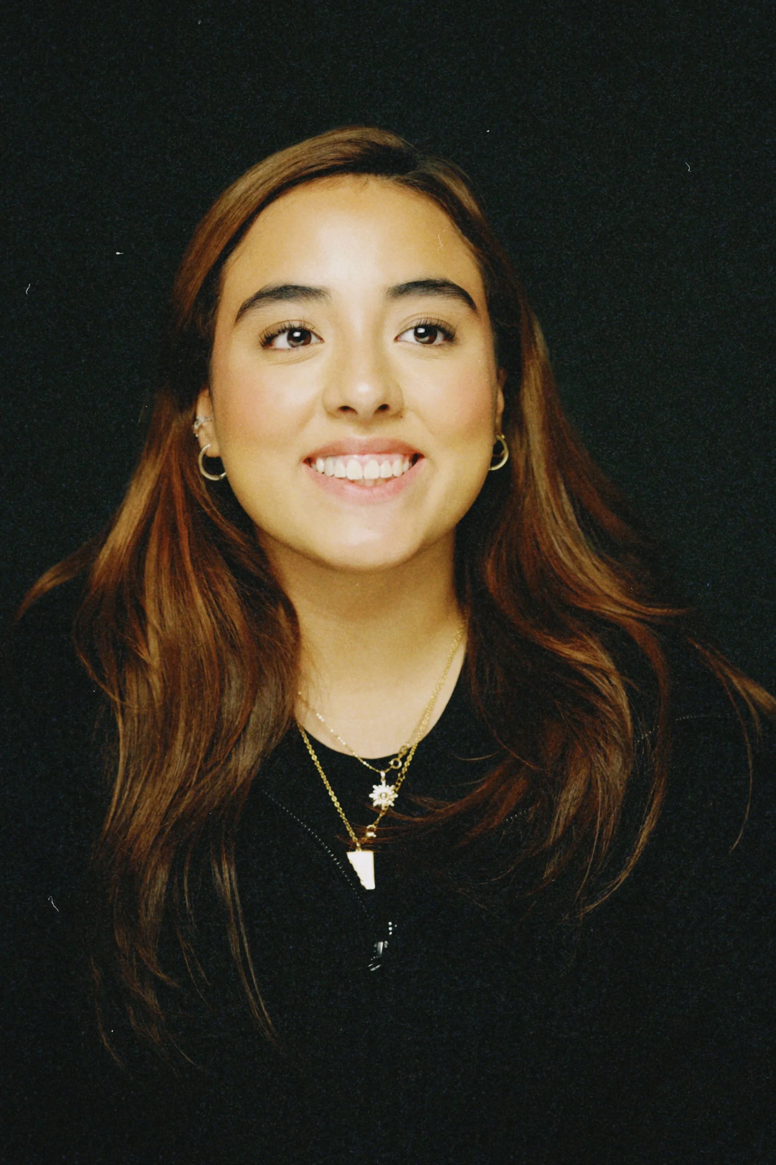 A young woman with long brown hair, smiling, wearing gold hoop earrings and layered necklaces, against a black background.