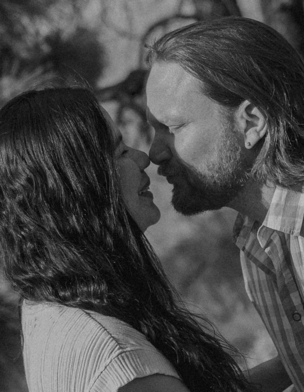 A black and white photo of a man and woman close enough to touch noses, with eyes closed, outdoors.