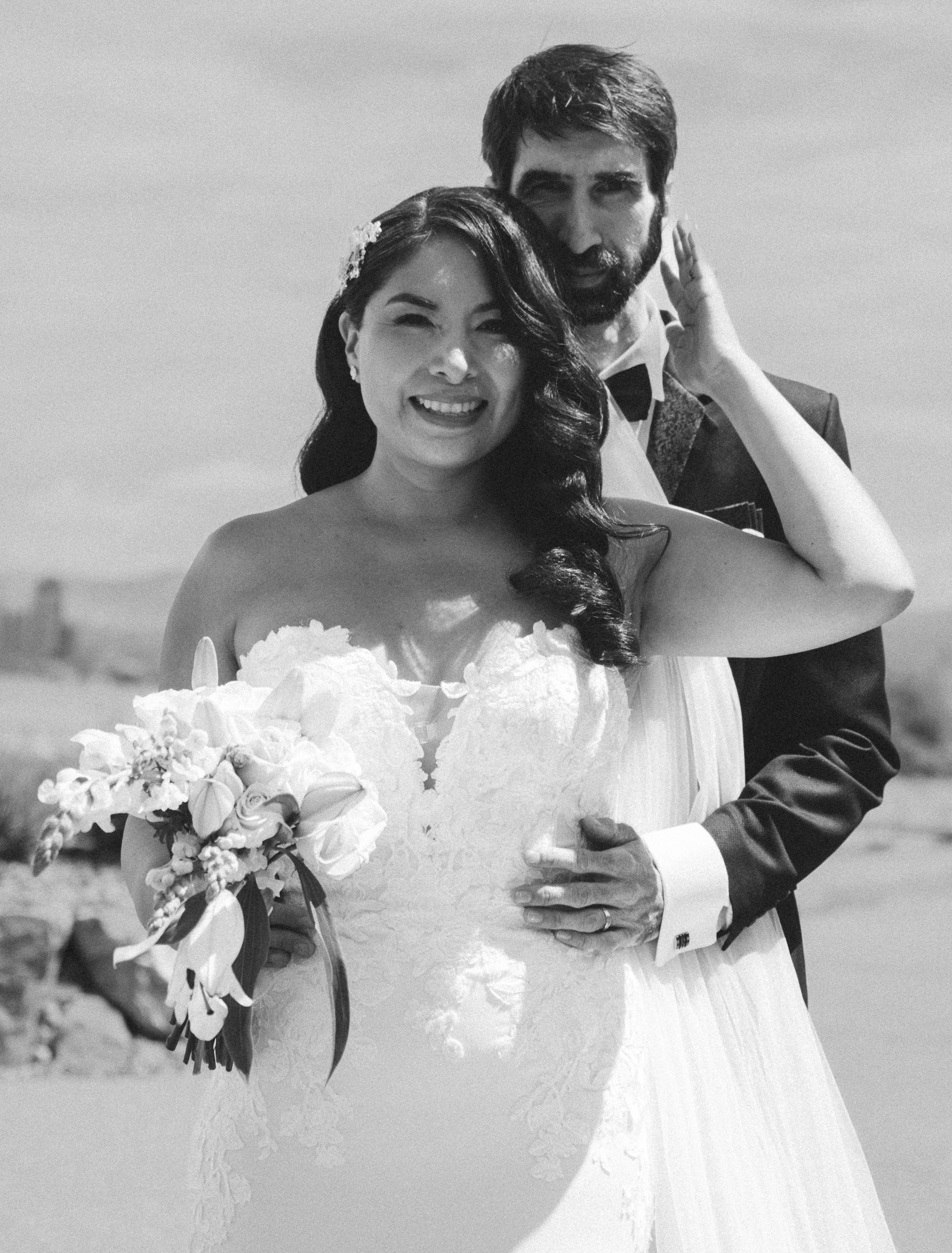 Black and white photo of a smiling bride in a strapless wedding dress holding a bouquet of flowers, standing close to a groom with a beard in a tuxedo, outdoors with a blurred background.