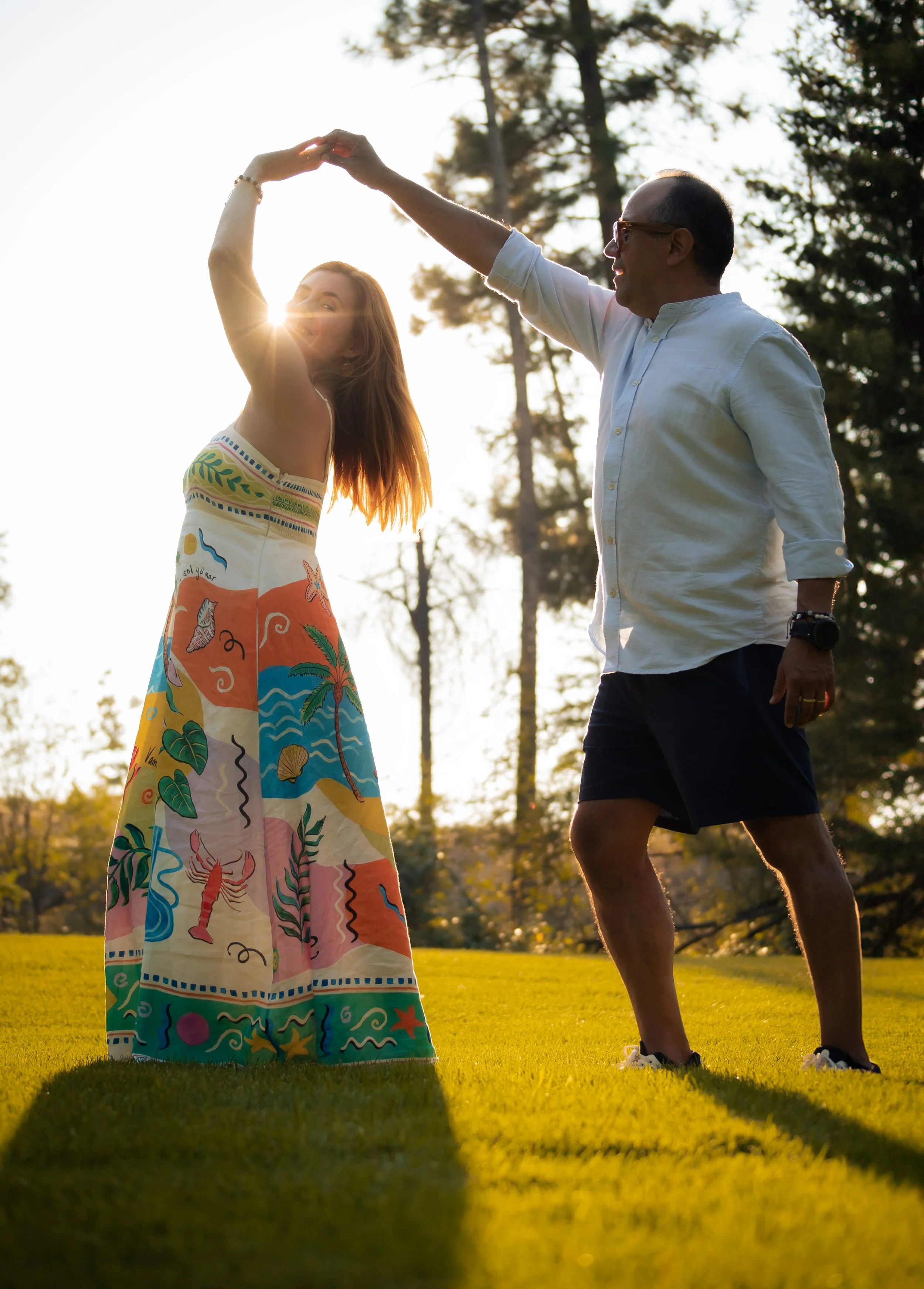 A man and woman dancing outdoors on a sunny day with trees in the background. The woman is wearing a colorful, patterned dress, and the man is wearing a white shirt and dark shorts.