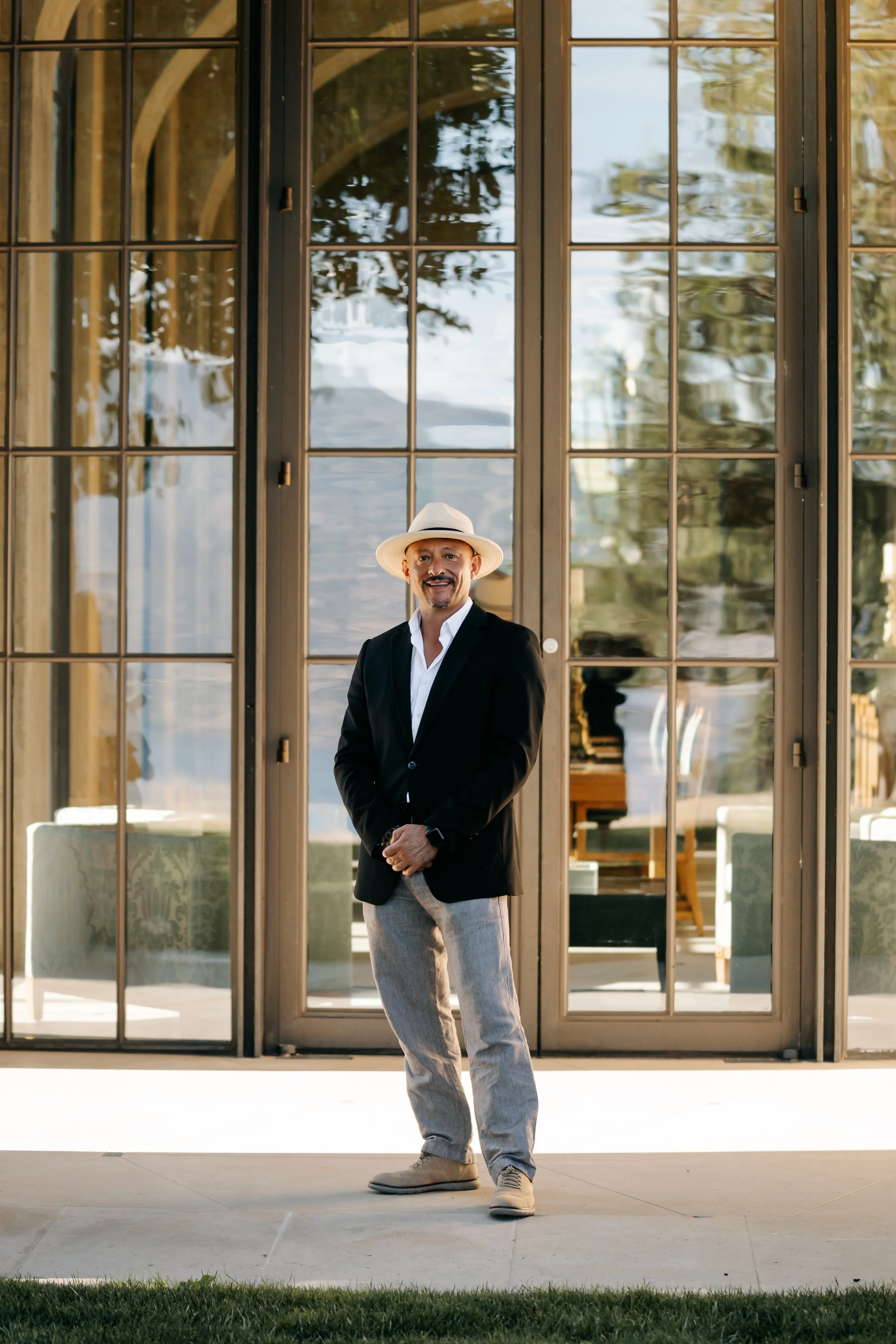 A man in a black blazer, white shirt, light gray pants, beige shoes, and a white hat standing outside in front of glass doors, smiling at the camera.