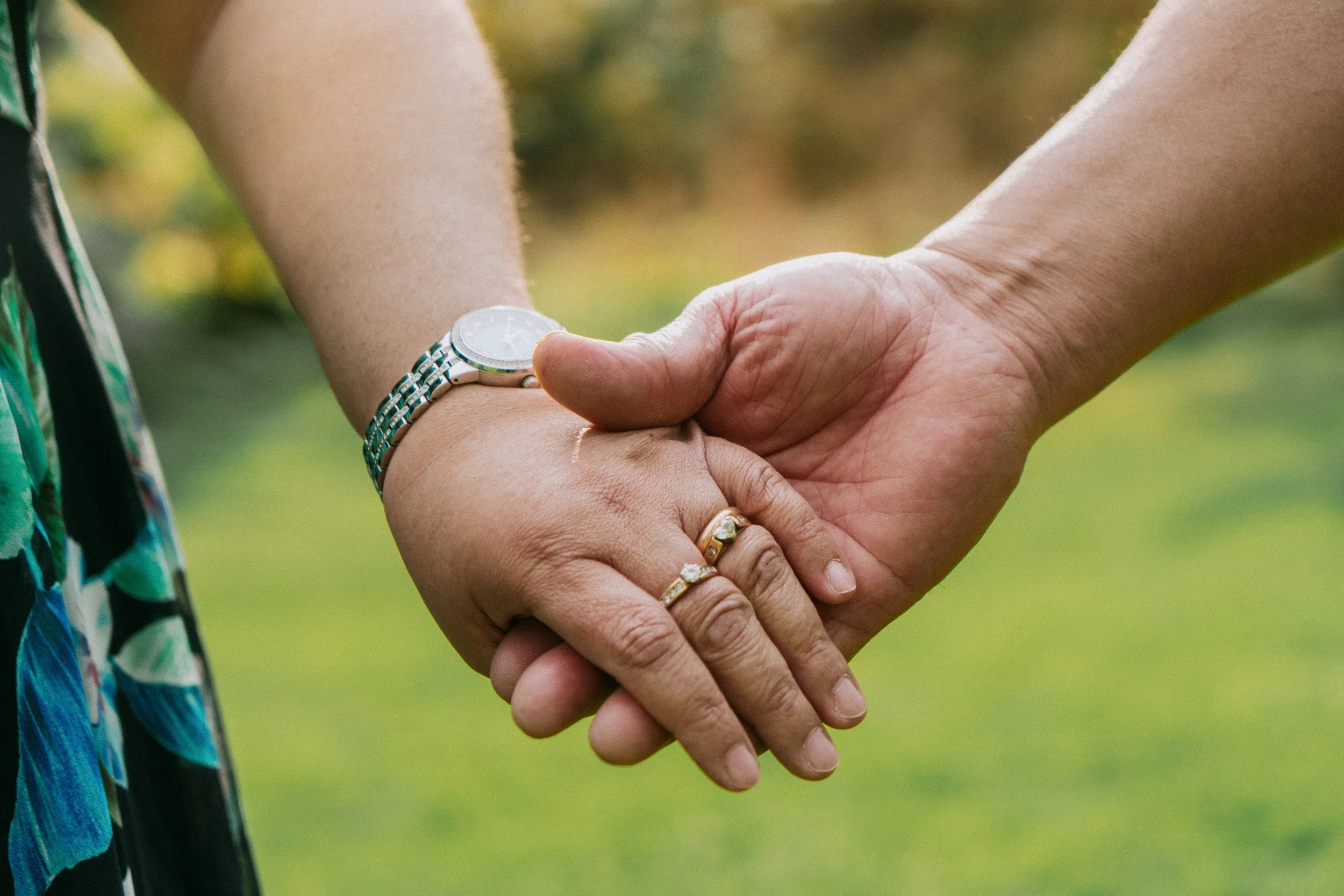 A couple holding hands outdoors, with one person wearing a ring and a wristwatch, the background is green and blurry.