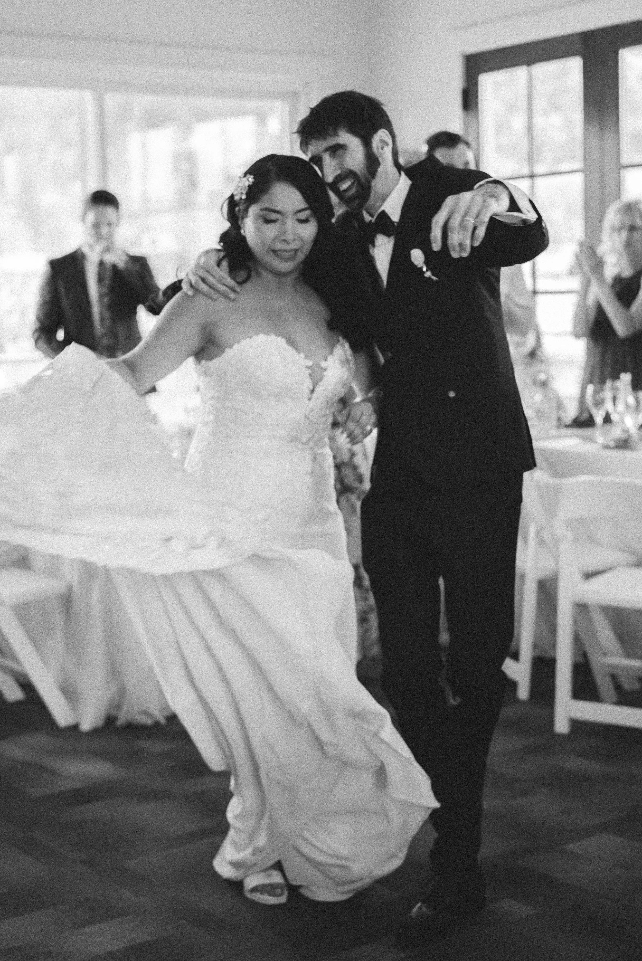 A couple, a bride and groom, dancing and smiling at their wedding reception with guests clapping in the background.