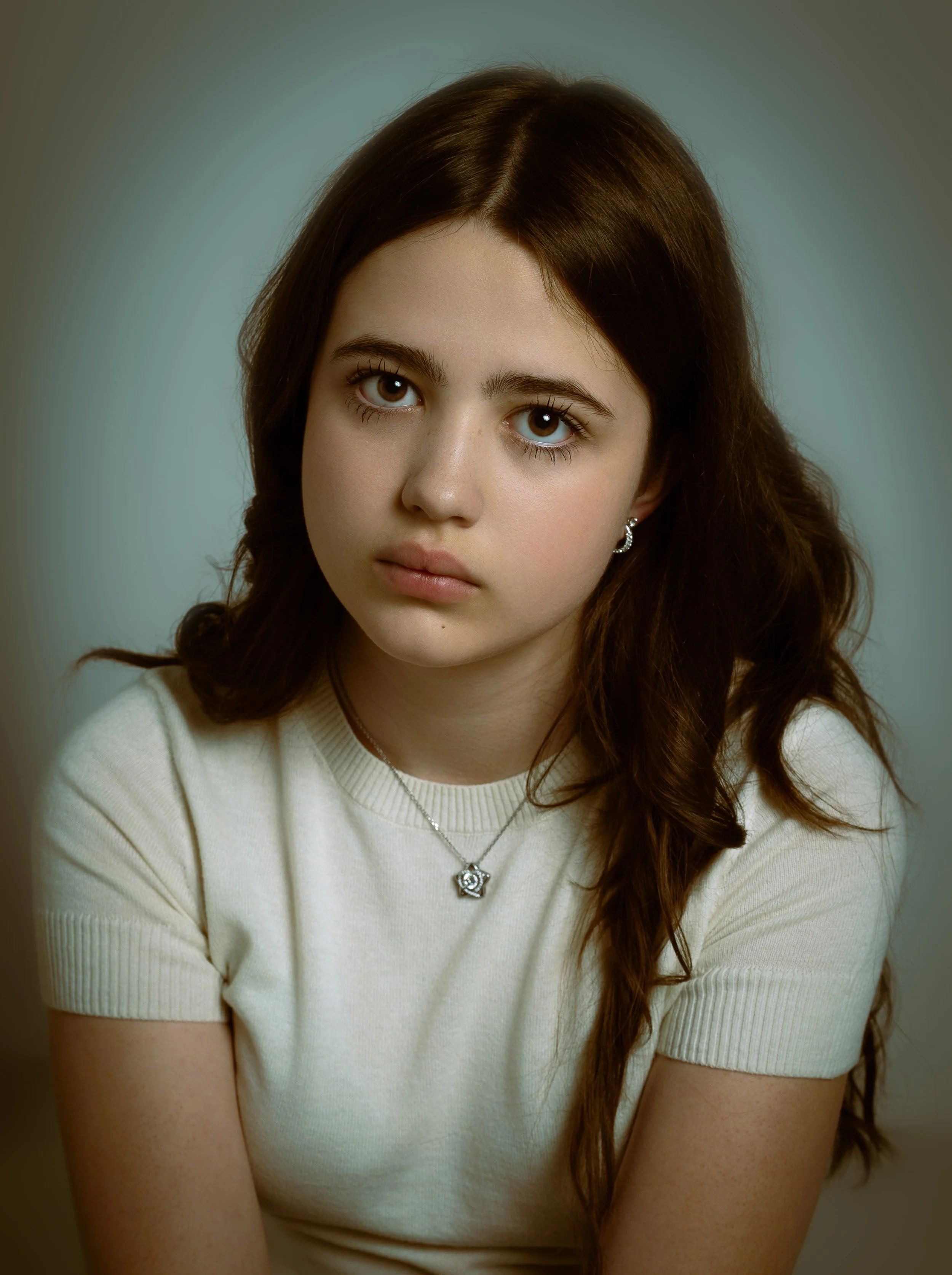 Young woman with long brown hair and gray eyes, wearing a cream-colored shirt and a star-shaped pendant necklace, looking directly at the camera with a neutral expression.