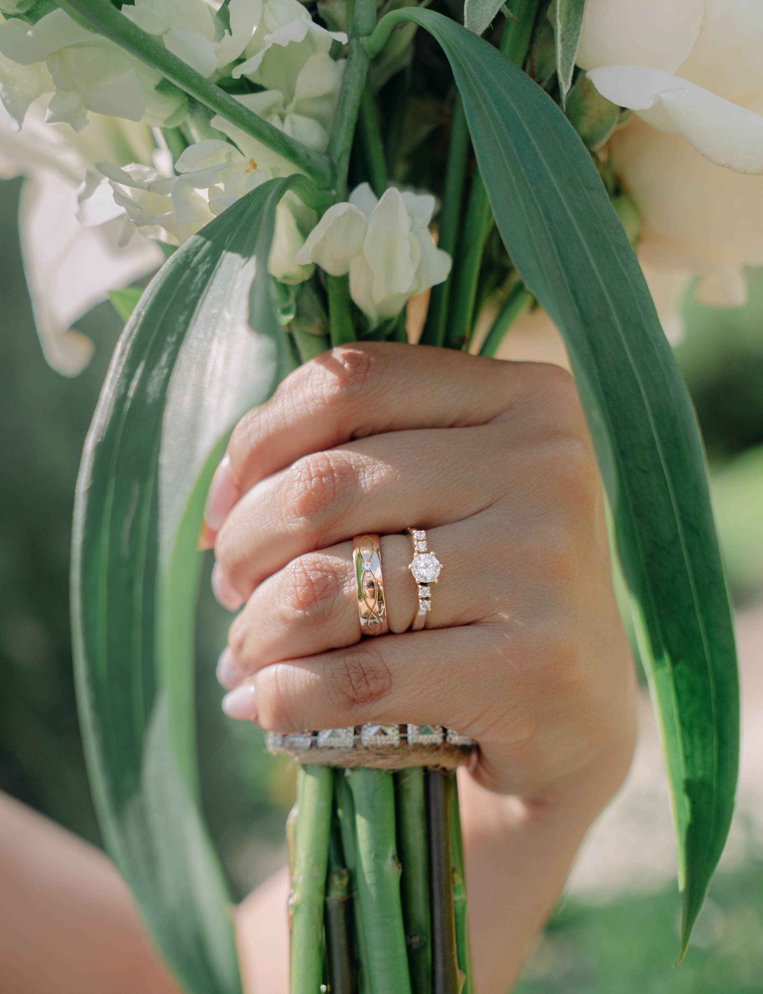 Hand holding a bouquet of white flowers with three rings on fingers, including a diamond engagement ring.