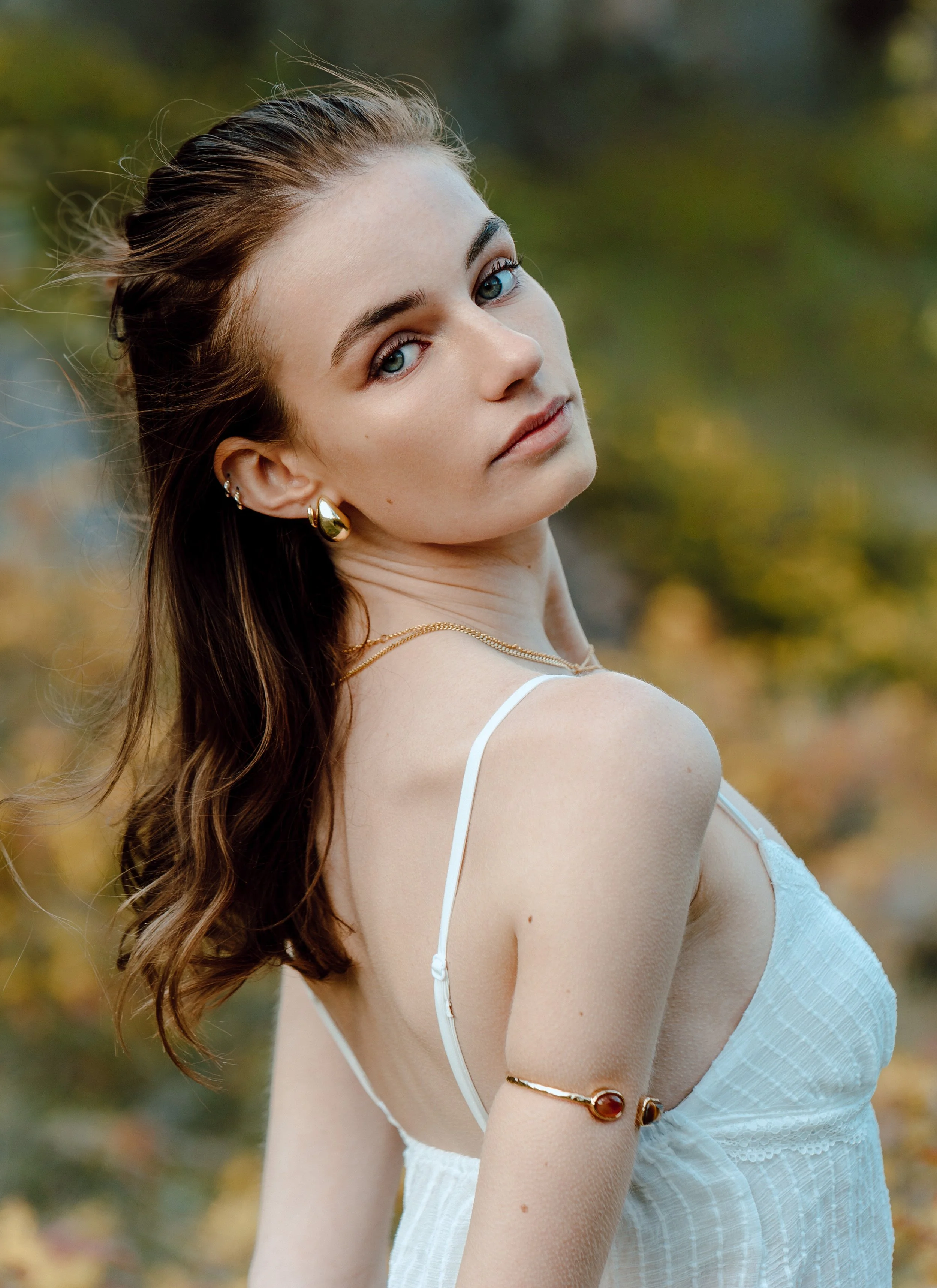 A young woman with brown hair and blue eyes, wearing gold jewelry, in a white dress outdoors with a blurred autumn background.