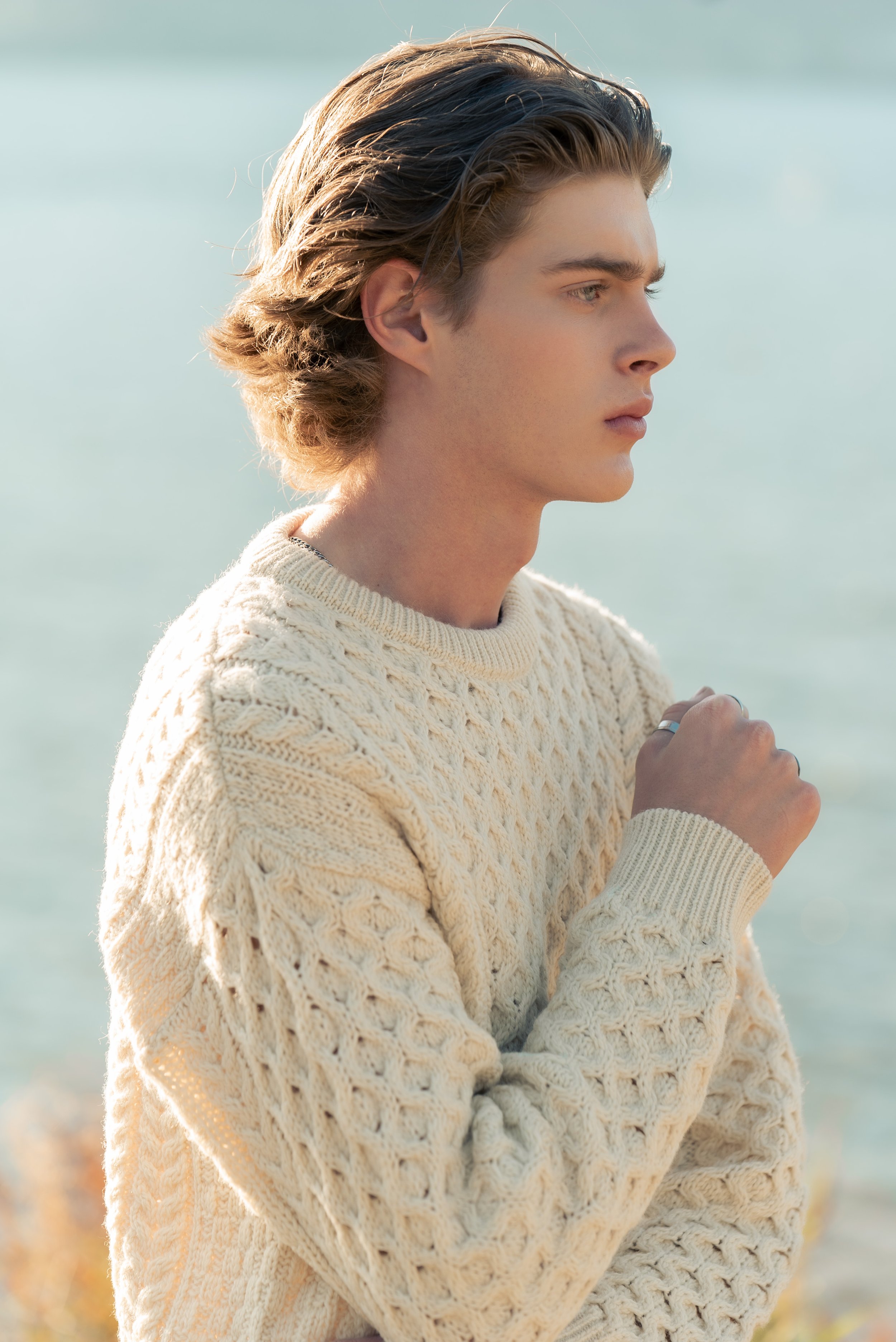 Young man with wavy hair looking thoughtfully at the water, wearing a cream-colored cable-knit sweater by the water.