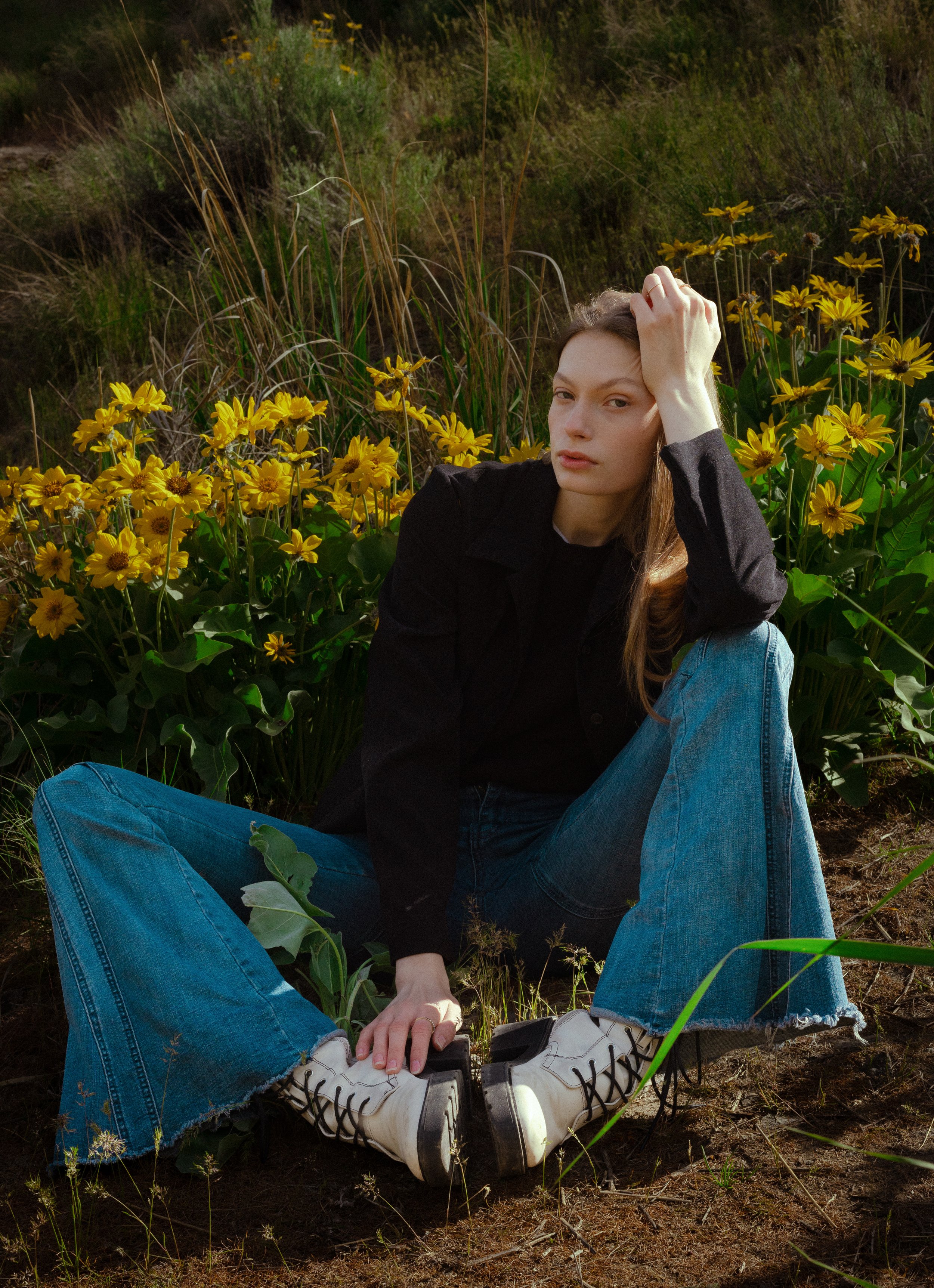 Young woman sitting on the ground amidst yellow flowers and greenery, with one hand on her forehead and wearing white boots, blue jeans, and a black top.