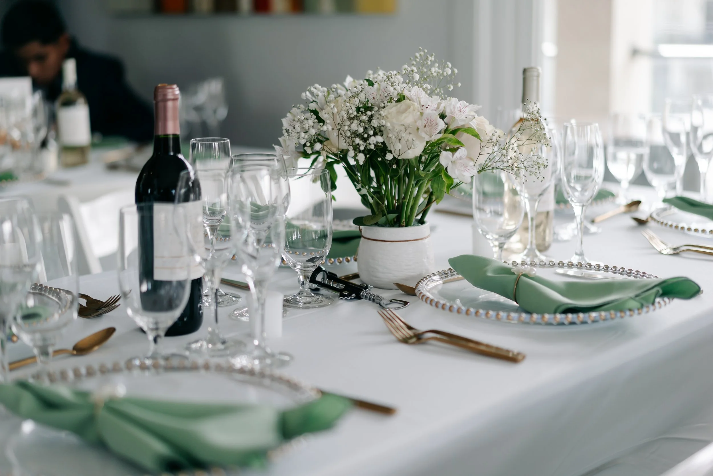 Elegant table setup with a white floral centerpiece, wine glasses, green napkins, and gold utensils, set for a formal event.