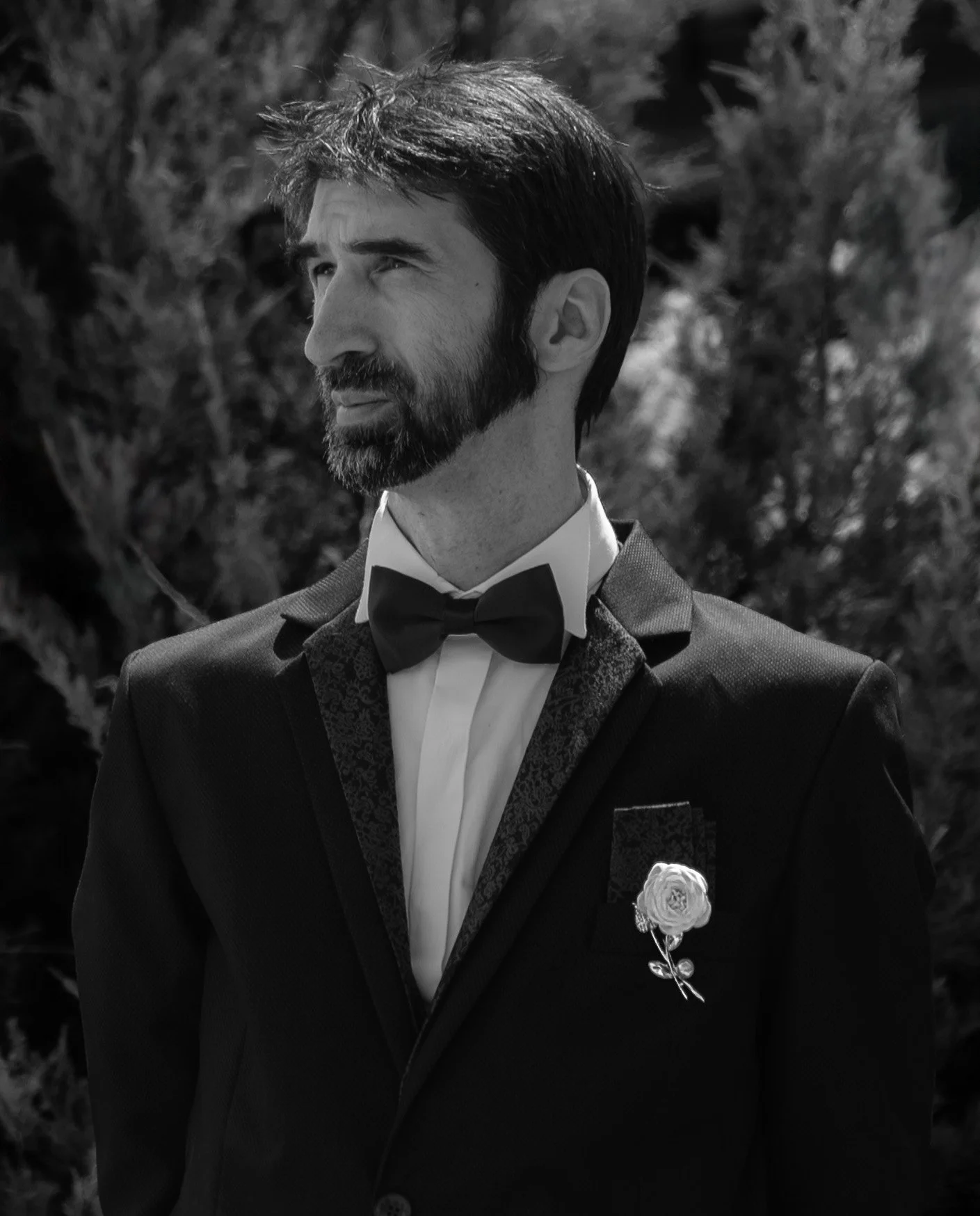 A man in formal attire, including a tuxedo with a paisley bow tie and a boutonniere, standing outdoors with trees in the background, looking to the side in black and white.