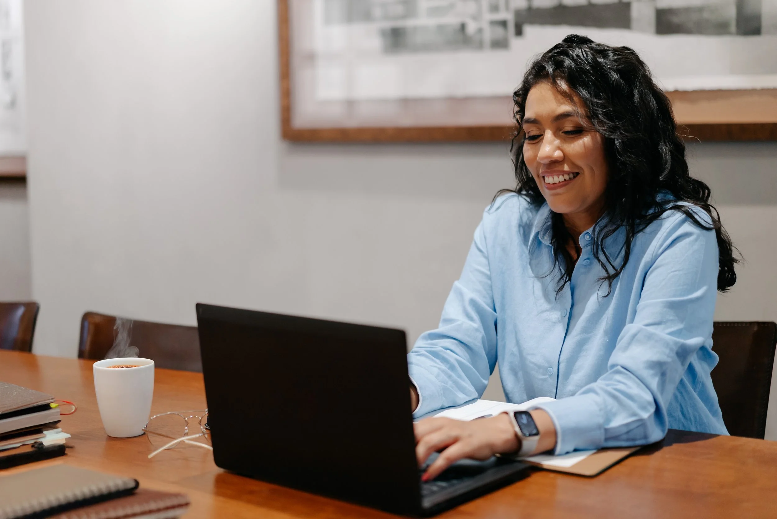 A woman with dark curly hair, wearing a light blue shirt and a smartwatch, is sitting at a wooden table working on a laptop. There is a steaming cup of coffee or tea, some notebooks, and a pair of glasses on the table.