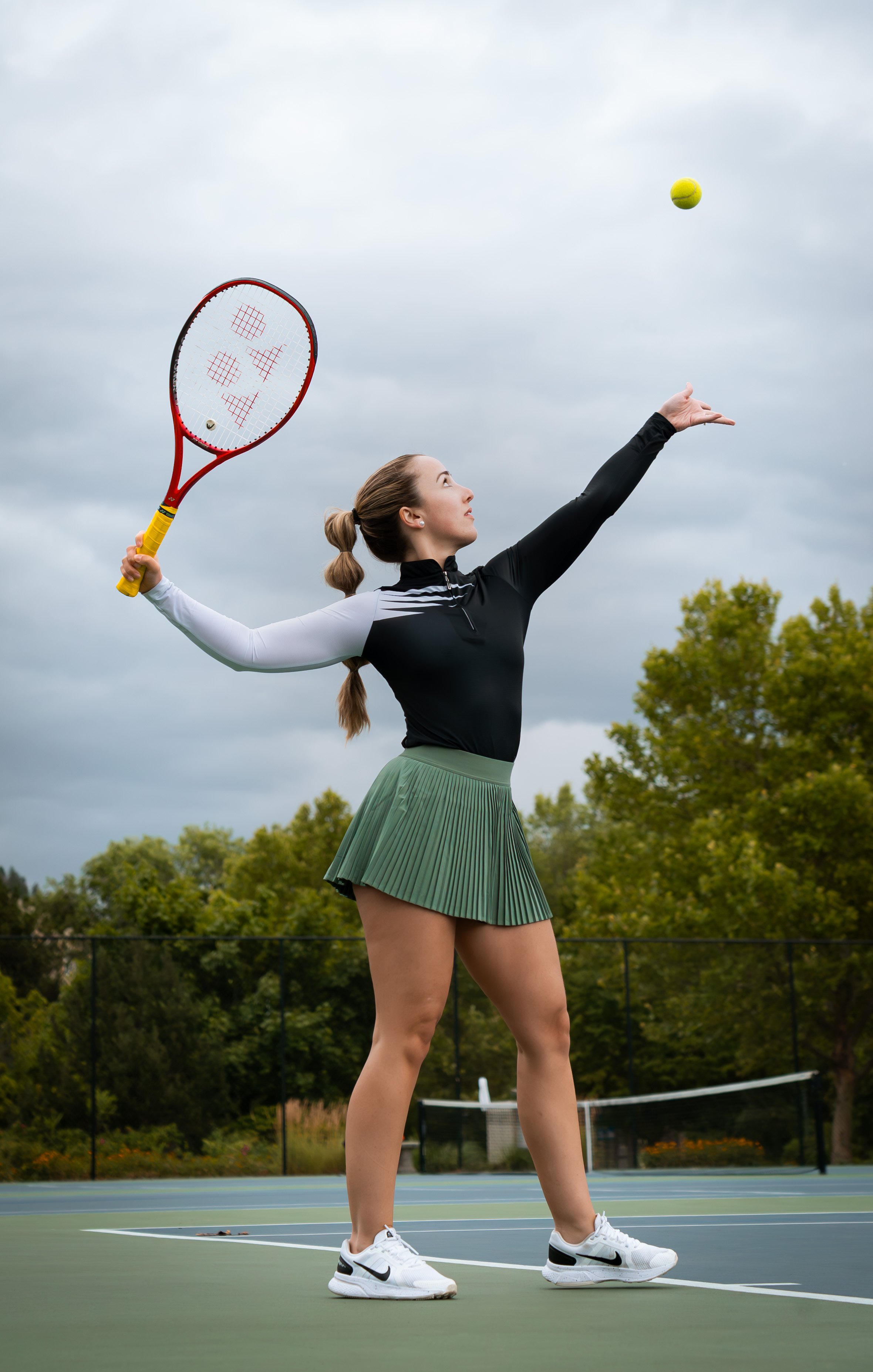 A woman on a tennis court preparing to hit a tennis ball with a racket, wearing a black and white sports top, a green pleated skirt, and white sneakers.