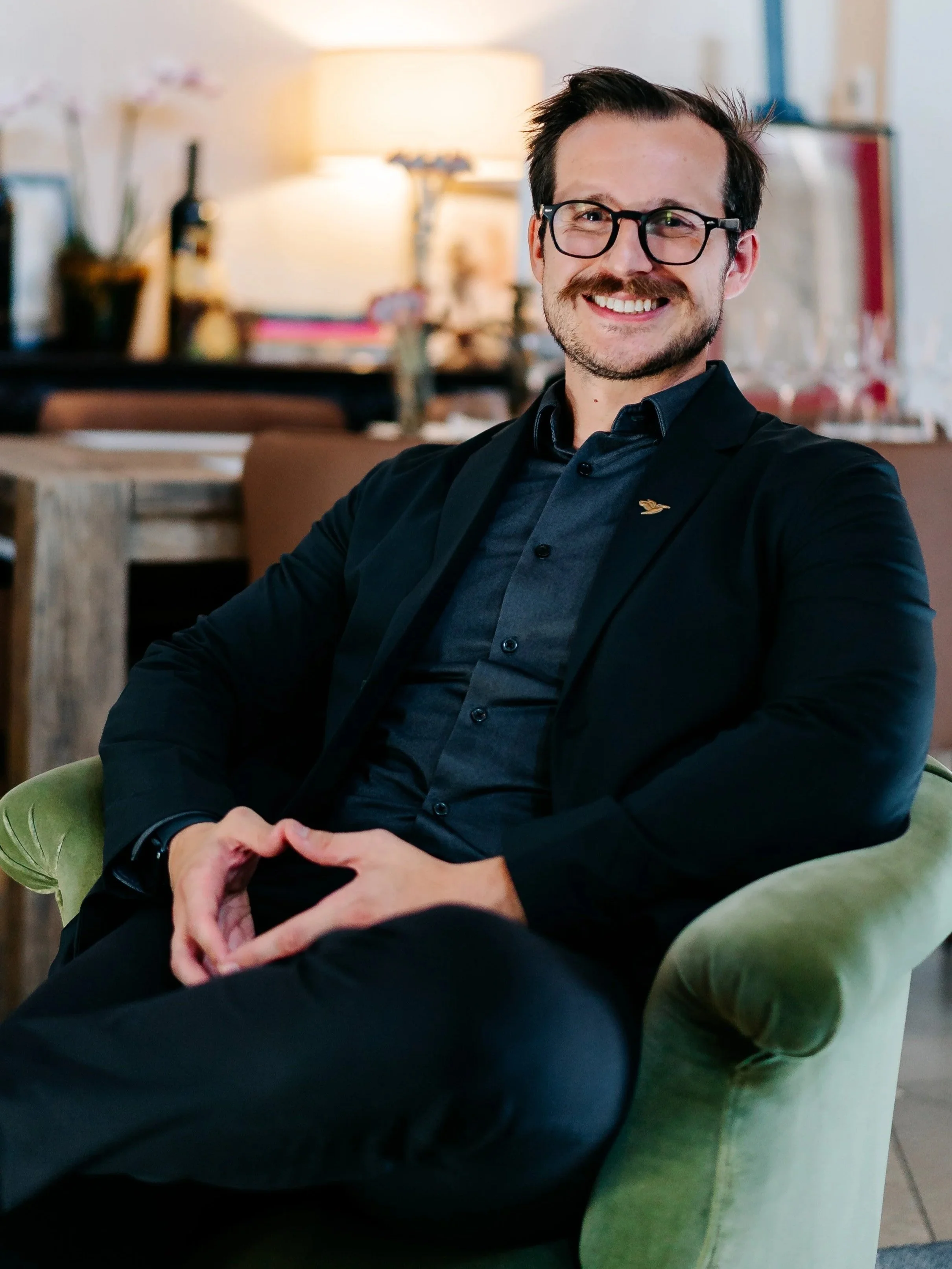 A man with glasses and a beard, smiling, sitting on a green armchair in a warmly lit room.