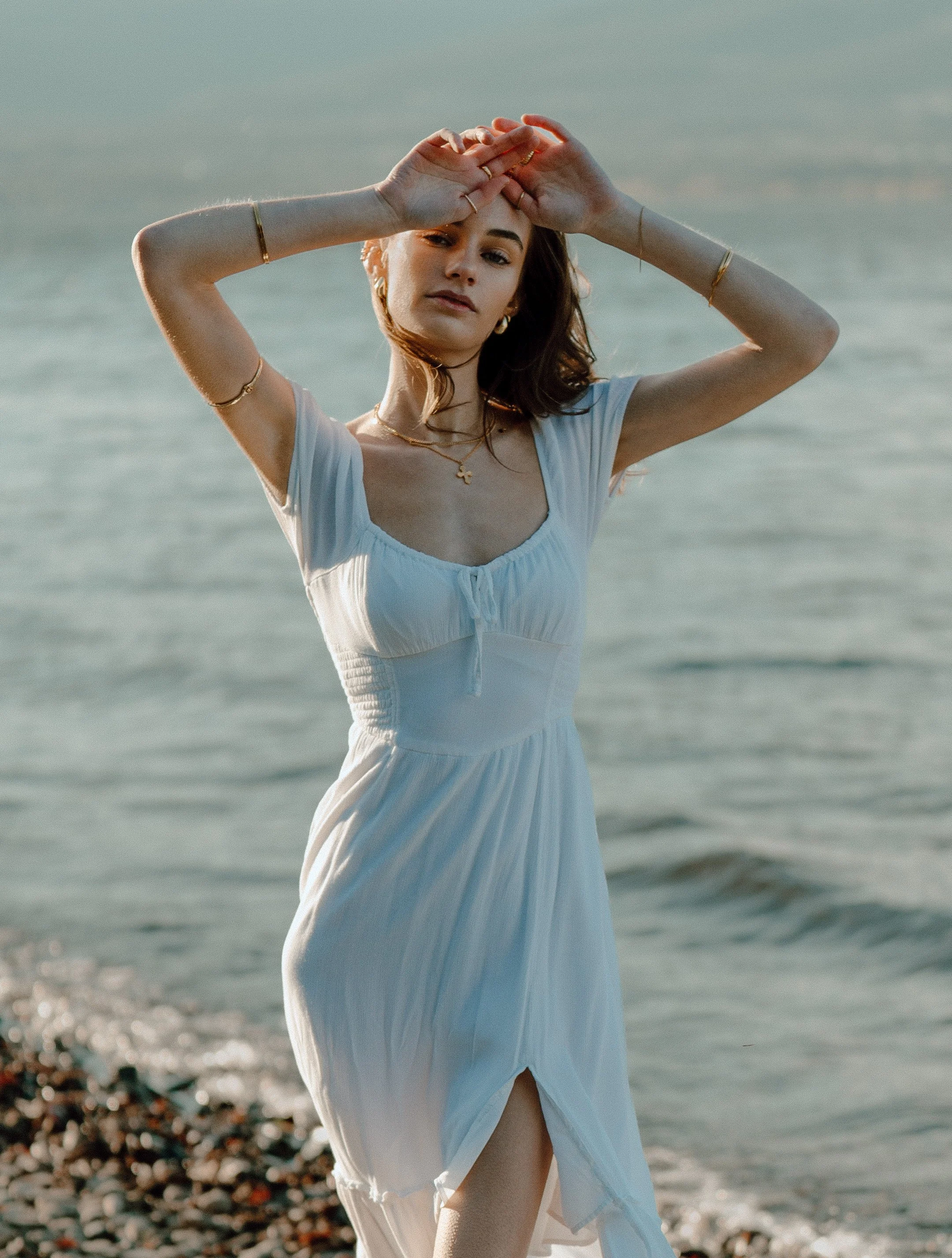 A woman in a white dress standing by the water, holding her hands above her head.