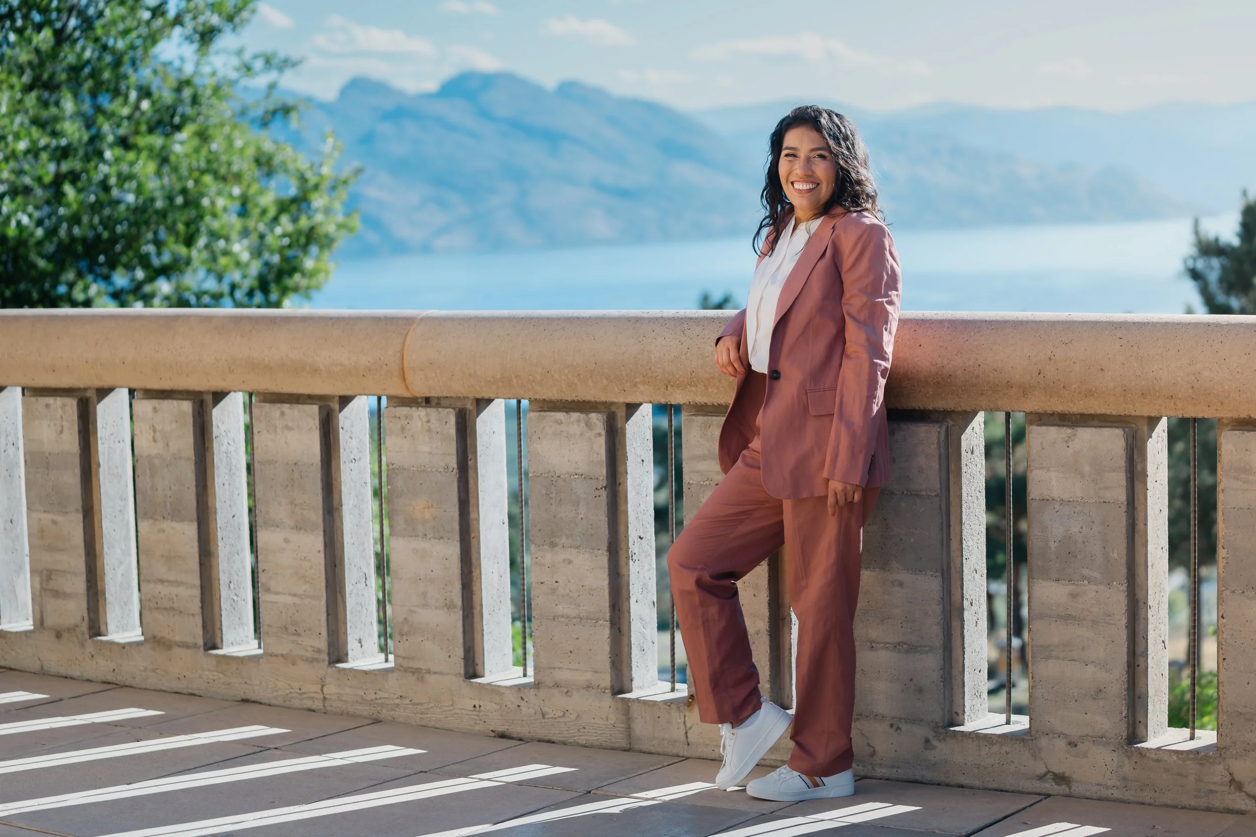 A woman in a pink pantsuit and white sneakers leaning against a stone railing outdoors, with water, trees, and mountains in the background, smiling at the camera.