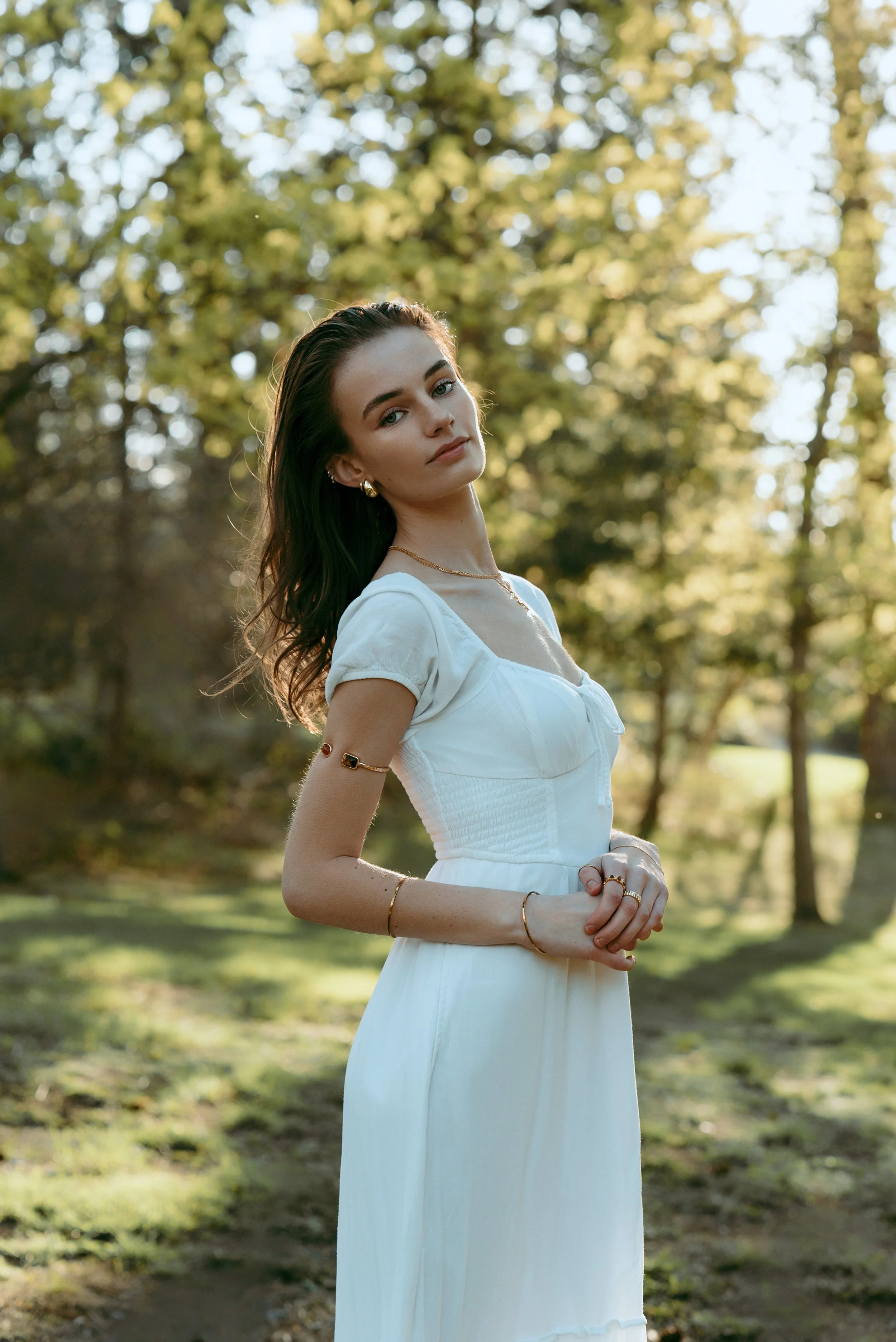 Young woman in a white dress standing outdoors in a wooded area during daytime with sunlight filtering through the trees.