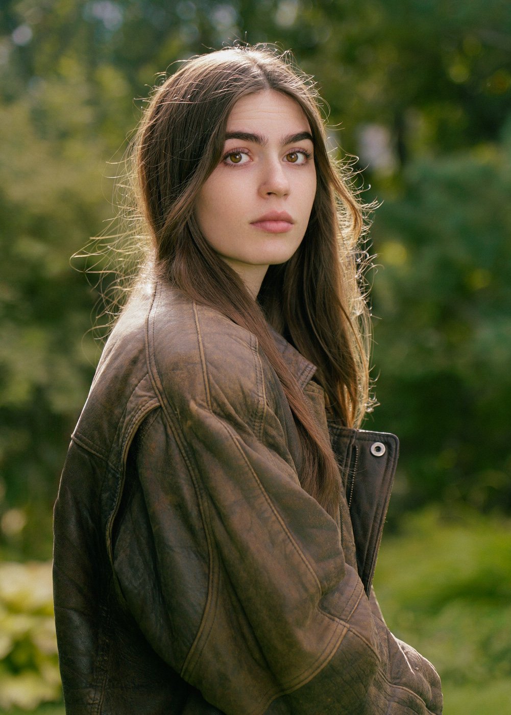 A young woman with long brown hair and fair skin is outdoors, wearing a brown leather jacket, with a green blurred background of trees and foliage.