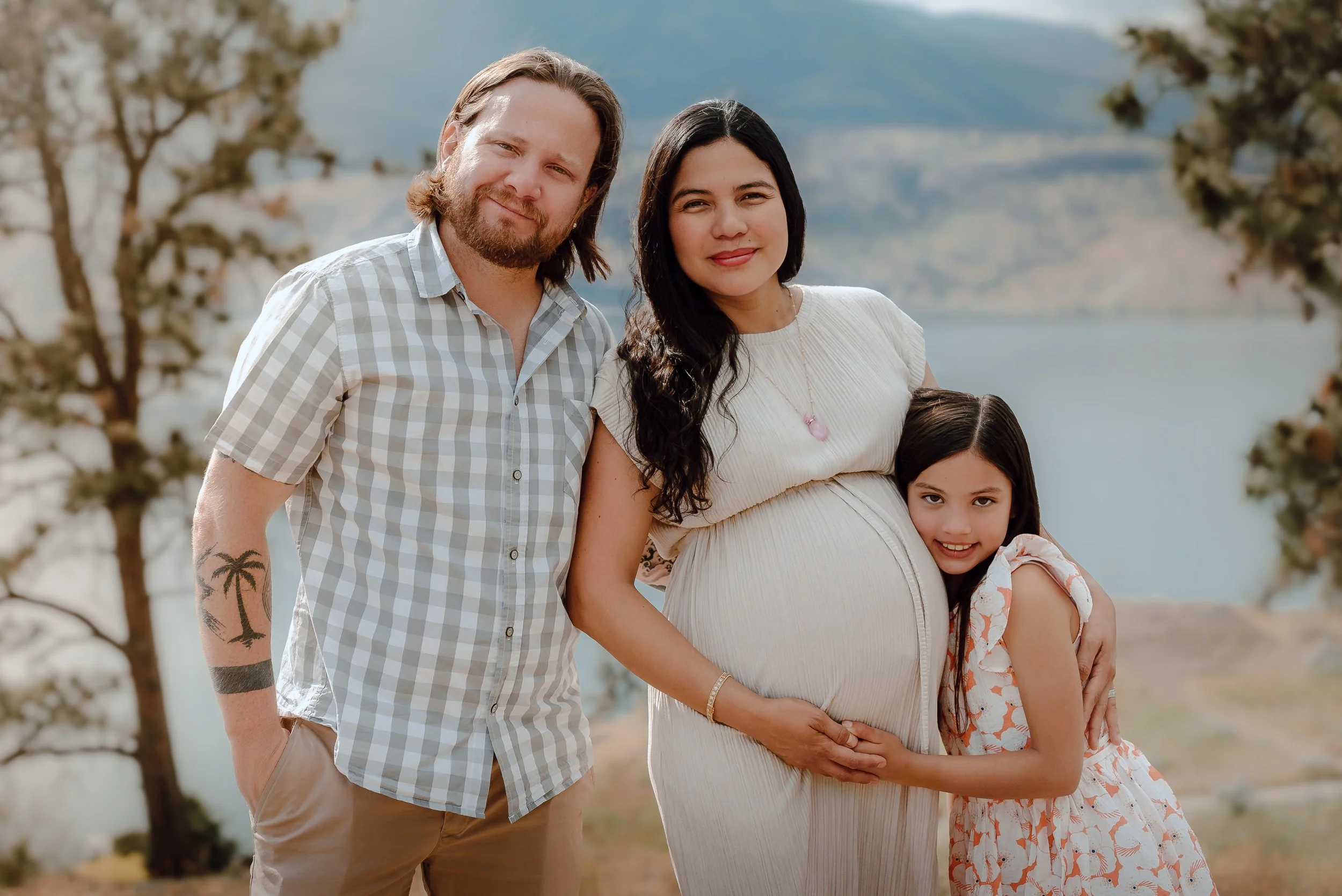 A happy family of three, including a pregnant woman, stands outdoors near a body of water with trees and mountains in the background. The woman is gently holding her belly, and a young girl is hugging her from the side. The man stands beside them with his arm around the woman’s shoulder.