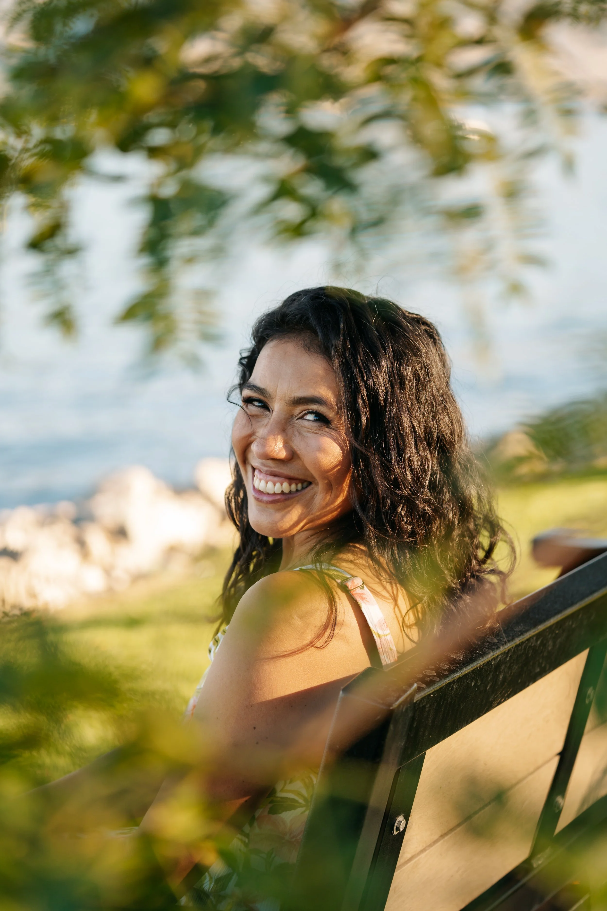 A woman with dark curly hair smiling and looking back over her shoulder while sitting on a park bench near water, with trees and rocks in the background.