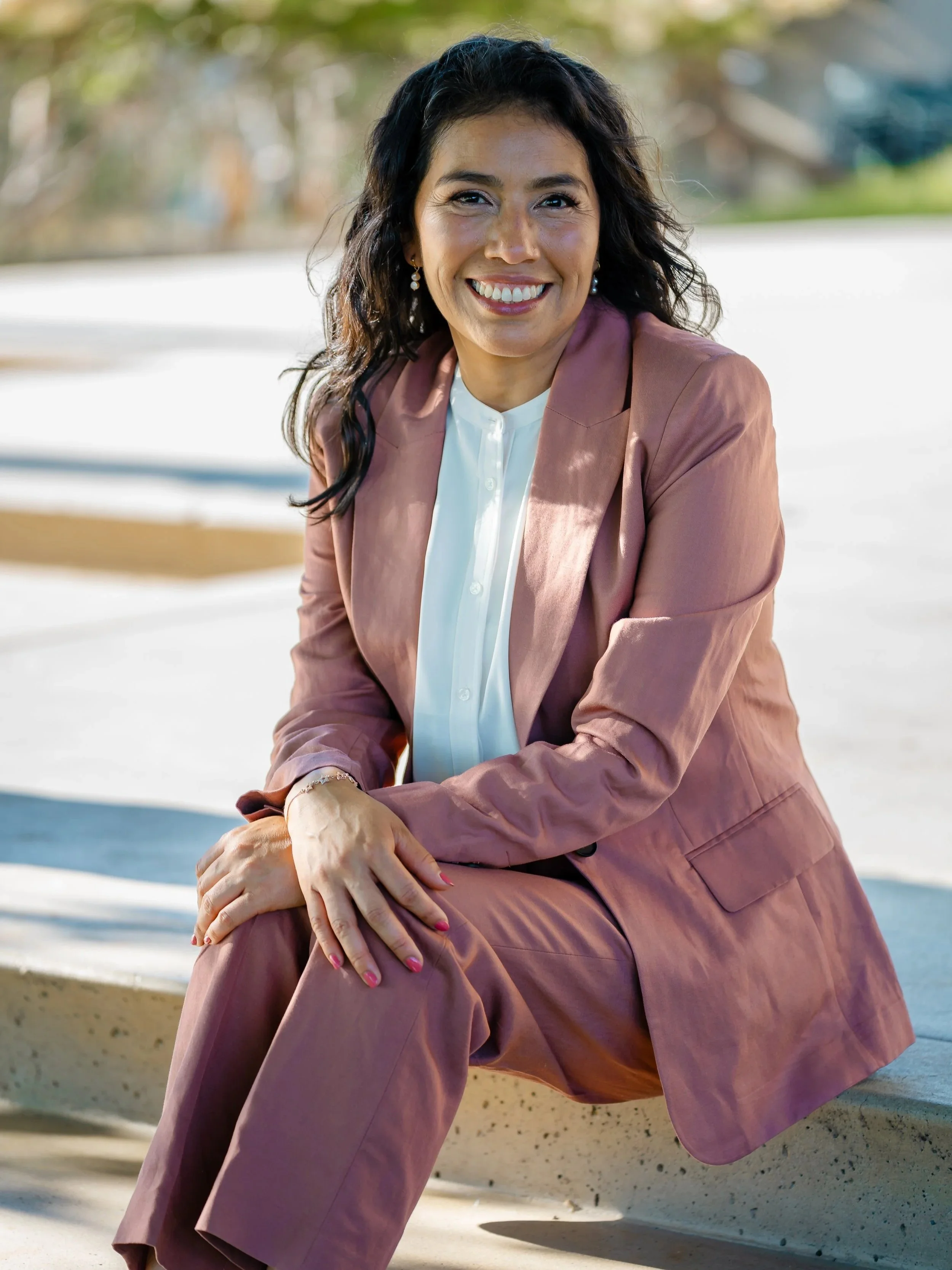 A woman with dark wavy hair, smiling, wearing a pink pantsuit and white shirt, sitting outdoors on a concrete bench on a sunny day.