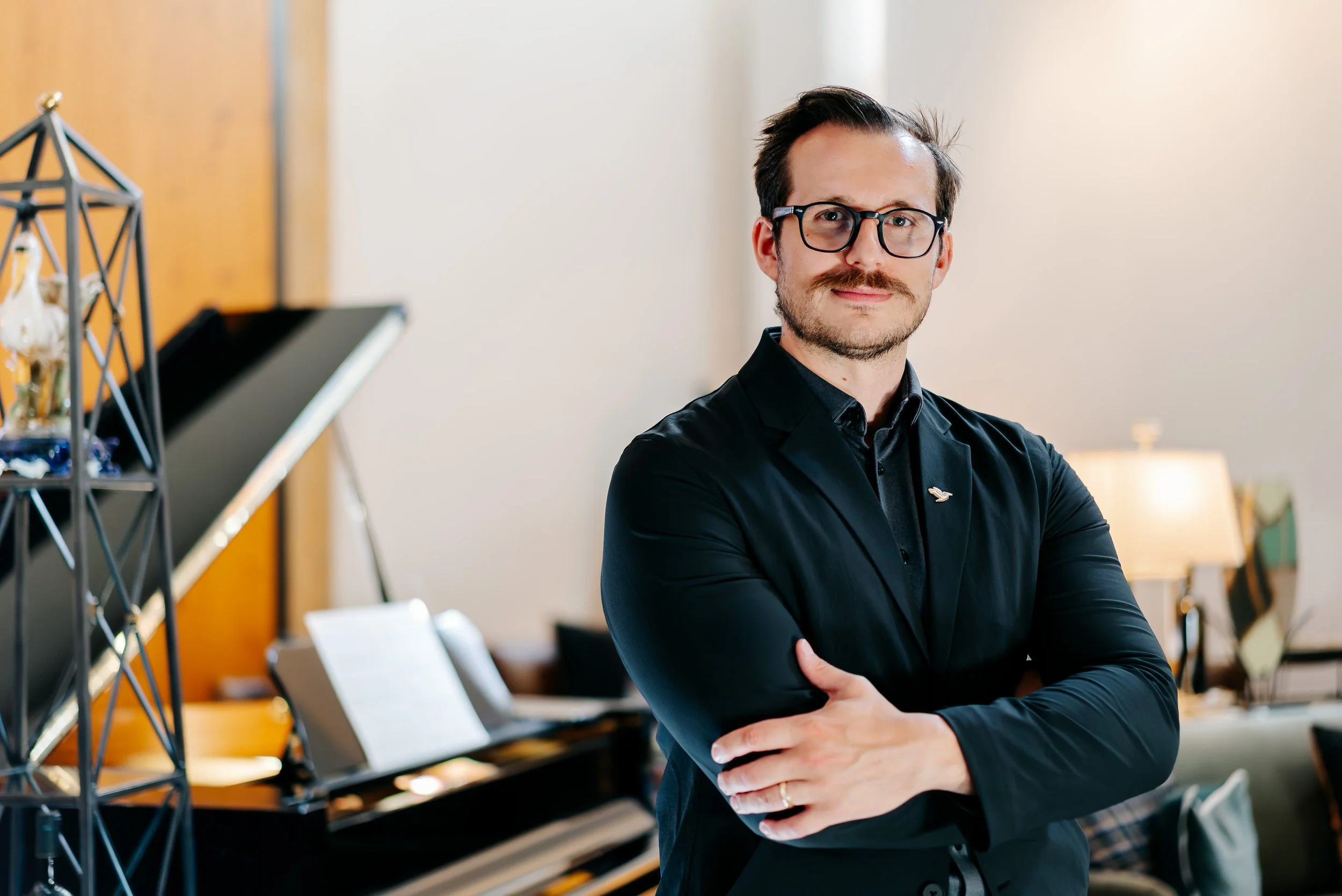 A man with dark hair, glasses, and a mustache standing with arms crossed in a room with a piano and music sheets in the background.