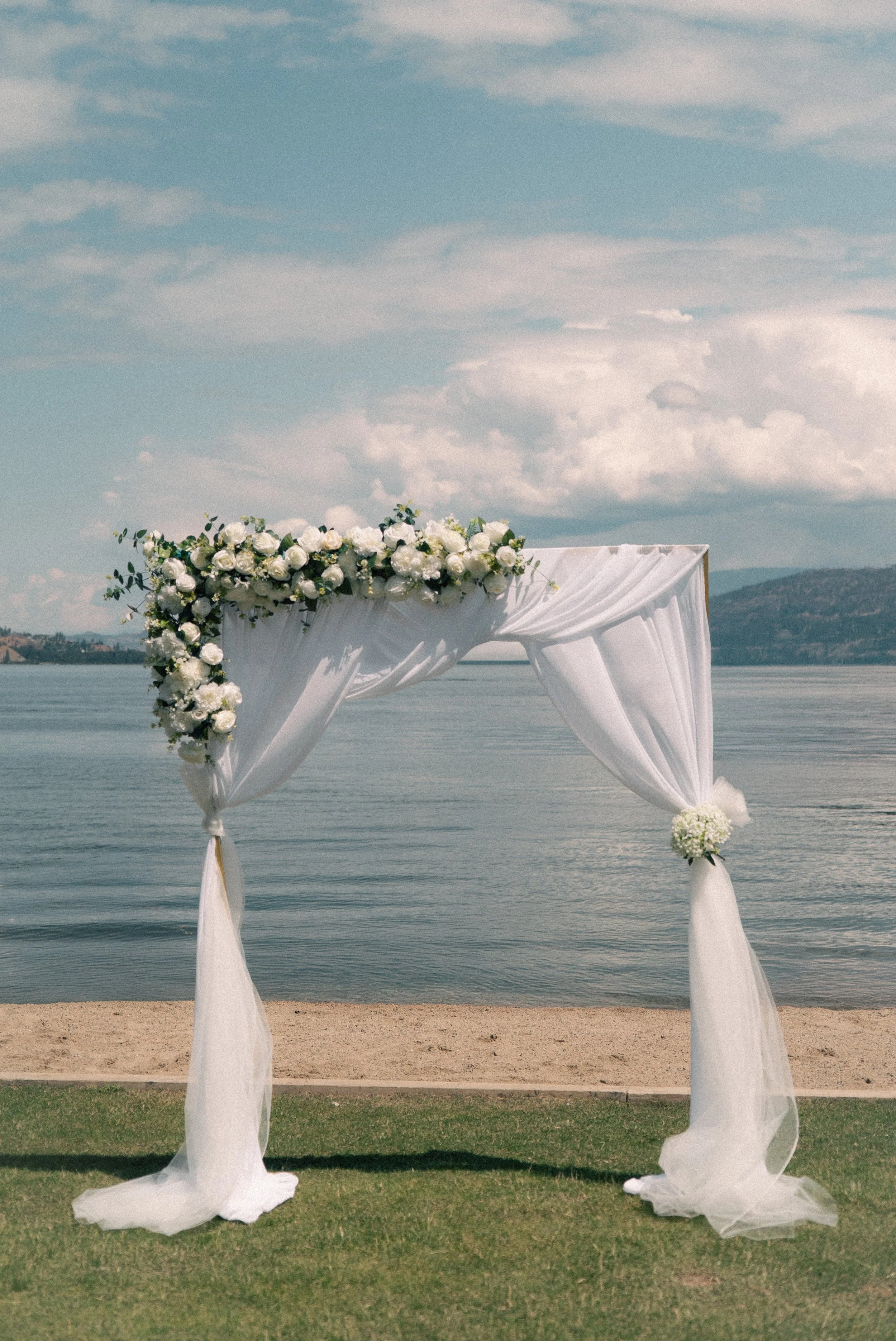 A decorated wedding arch with white drapes and white flowers, set on a grassy area by a lake with mountains in the background.