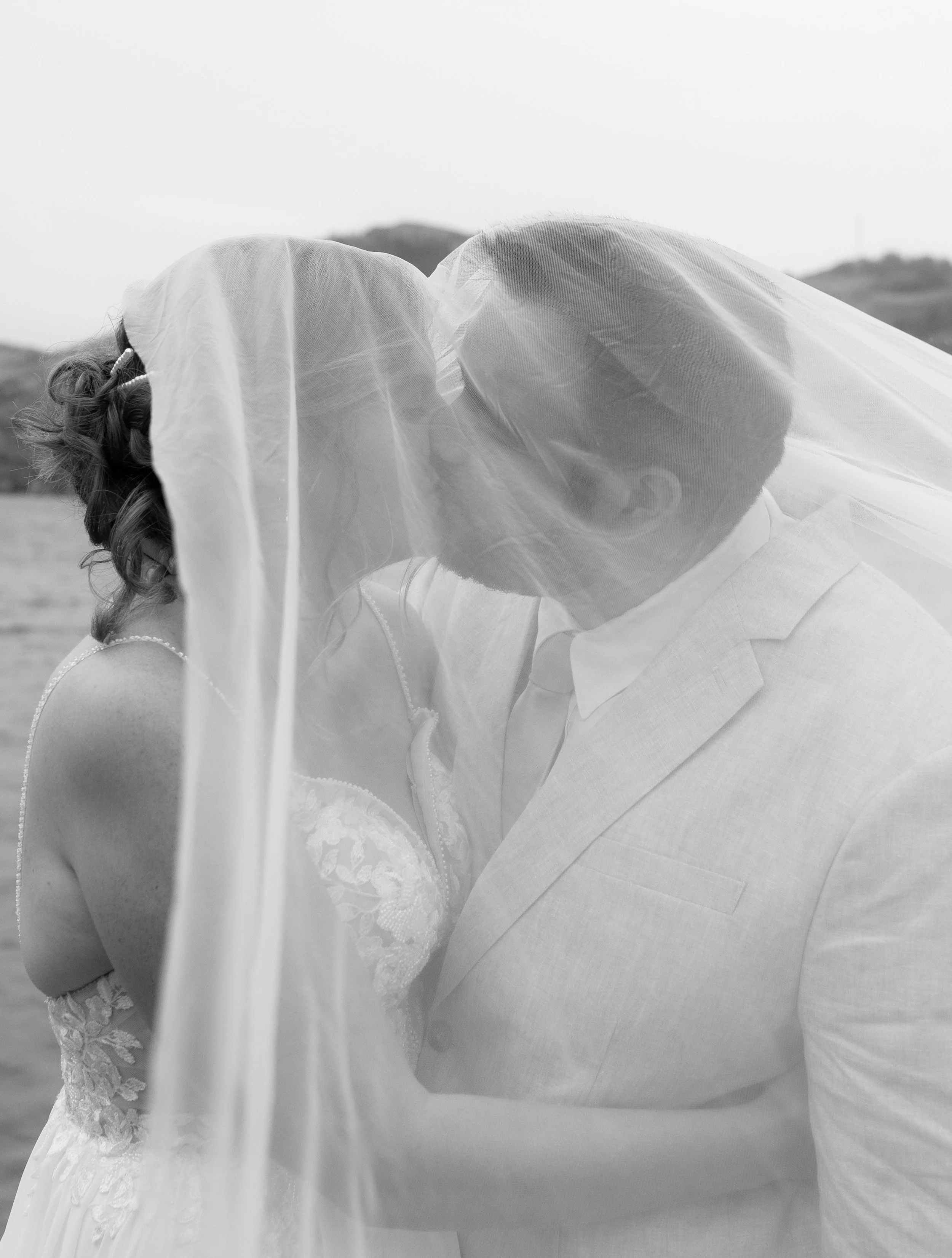 A black-and-white photo of a bride and groom sharing a kiss outdoors, with a veil covering part of their faces.