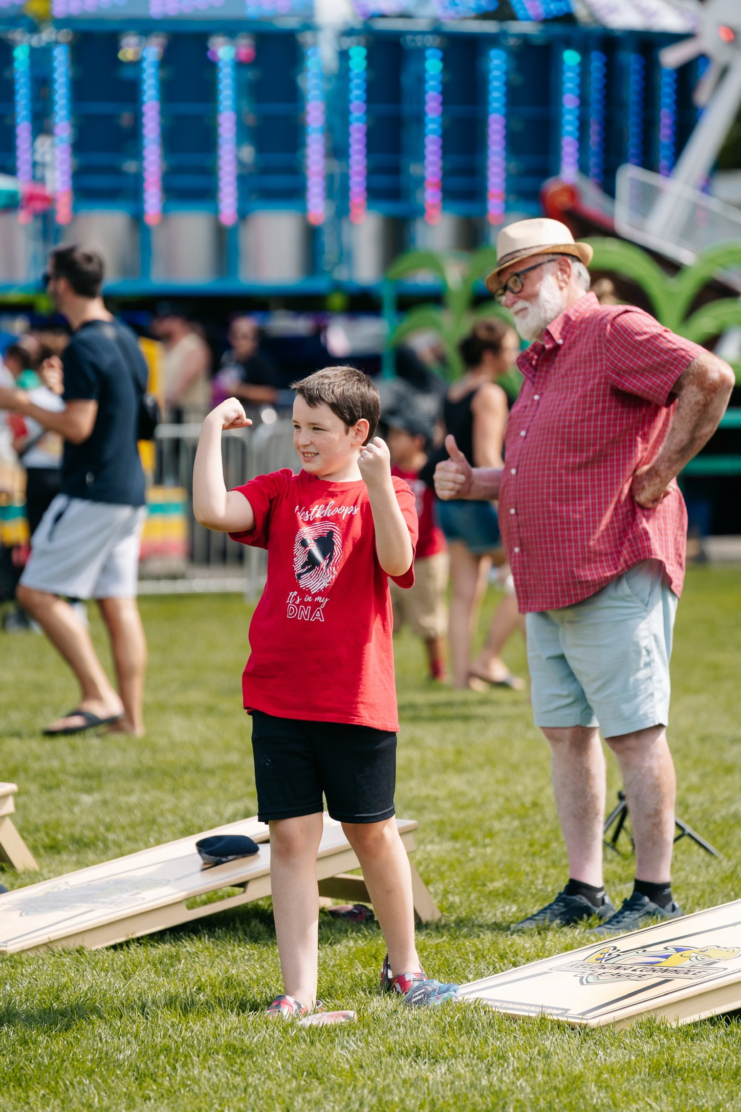 A young boy in a red shirt flexing his arms while an elderly man in a red checkered shirt and hat stands beside him giving a thumbs-up at an outdoor fairground.
