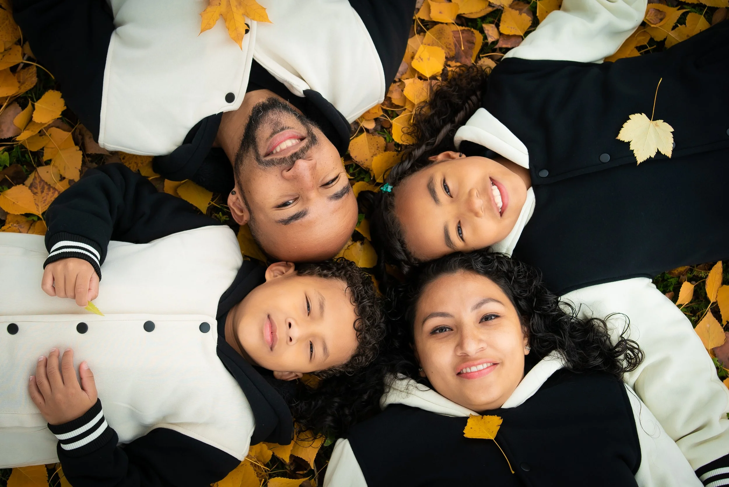 A family of four lying on the ground covered with autumn leaves, smiling and looking at the camera