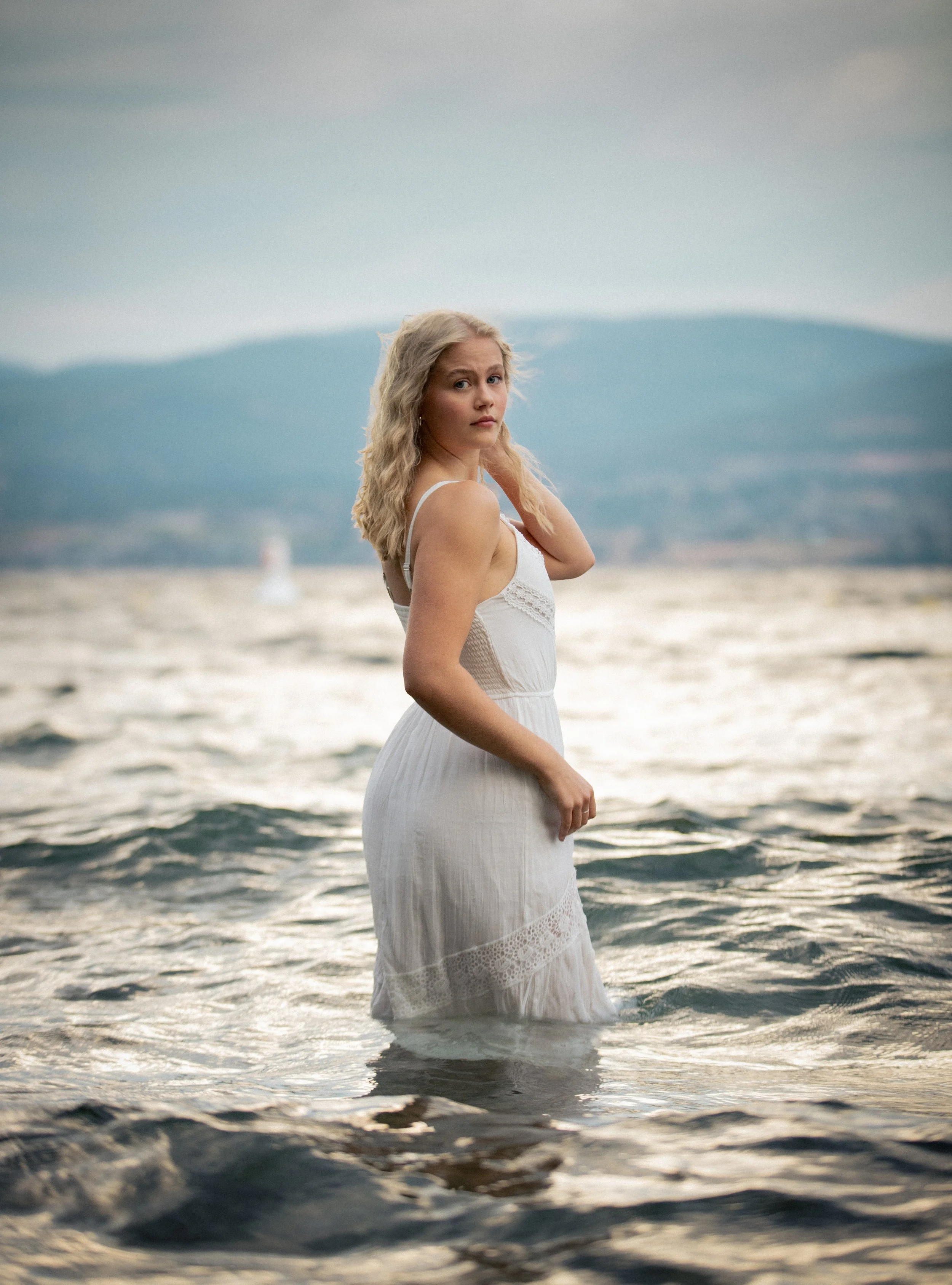 A woman in a white dress standing in the water at a beach during sunset or sunrise, with mountains in the background