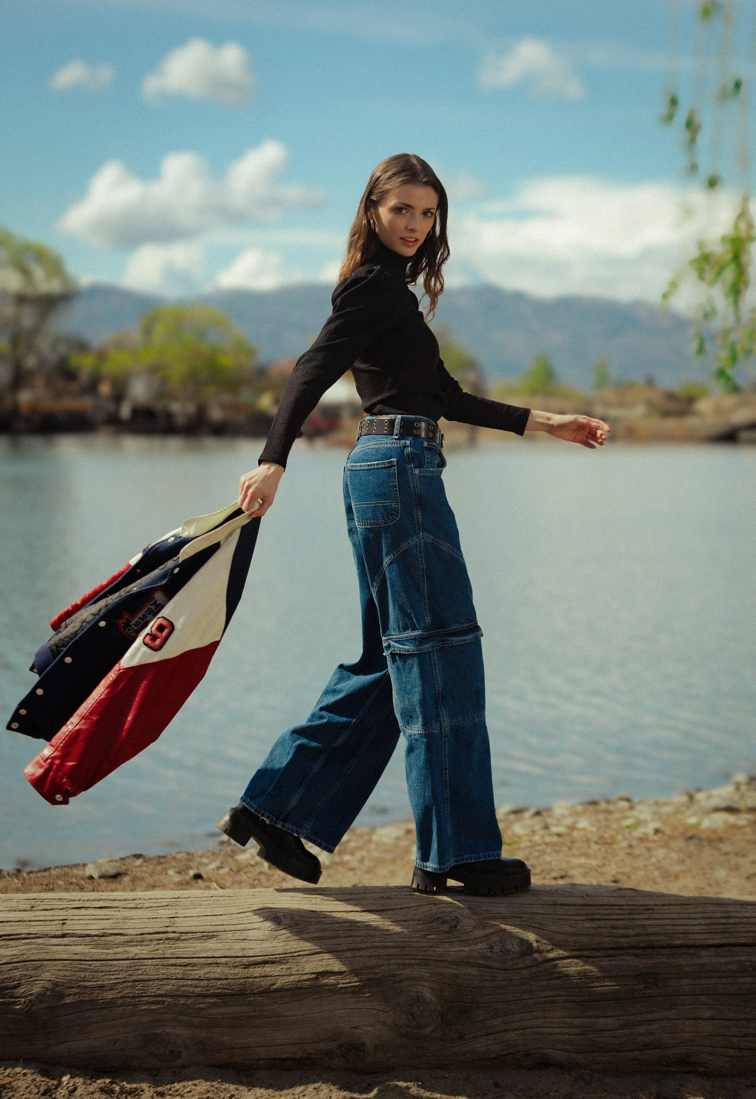 A woman walking along a lakeshore, holding a jacket with sports patches, dressed in a black top, wide-leg jeans, and chunky boots, with a scenic mountain and lake background.