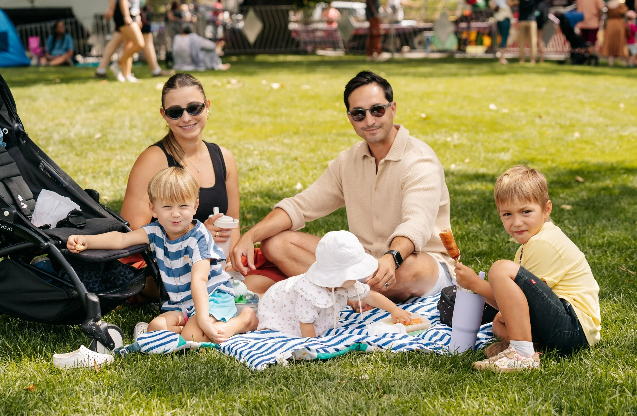A family of five having a picnic on the grass at a park. The family includes two adults and three children, sitting on a blue and white striped blanket. The woman is wearing sunglasses and a black top, the man is wearing sunglasses and a beige sweater. The children are sitting around them, one holding a skewer, another reaching towards a small toy, and one sitting in a stroller. In the background, other people are sitting and walking in the park.