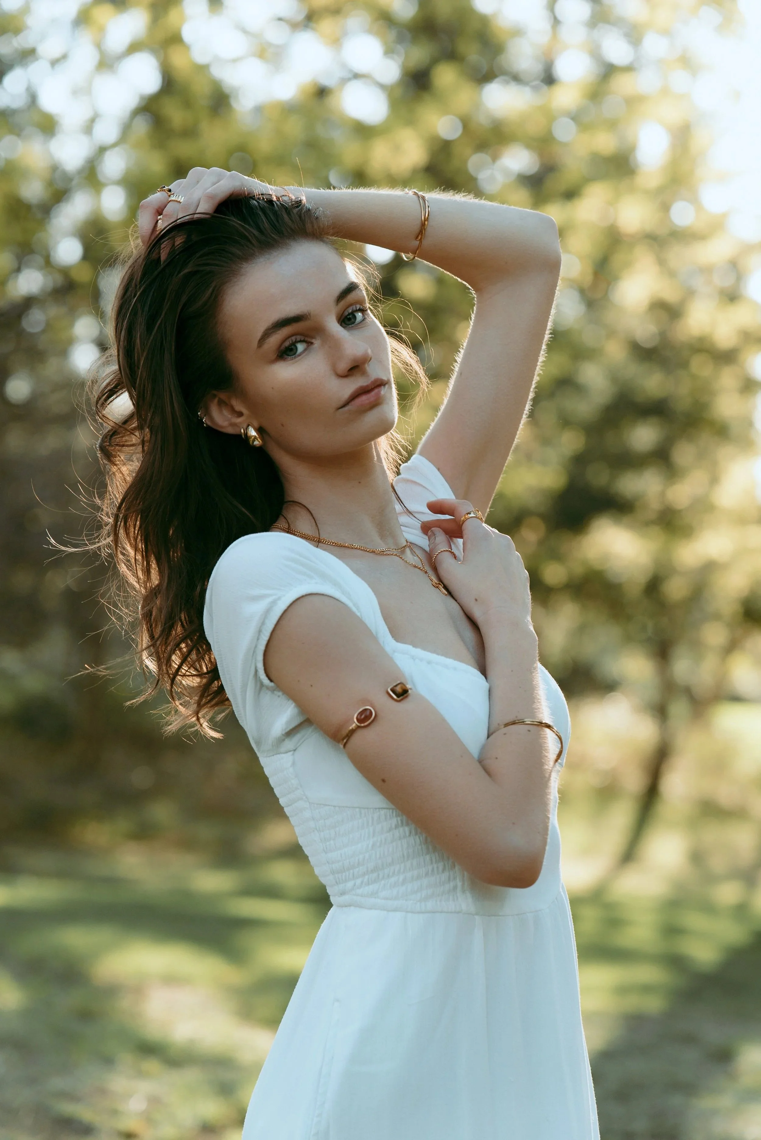 Young woman in a white dress standing outdoors, touching her hair with one hand and resting the other on her chest, surrounded by blurred trees and sunlight.