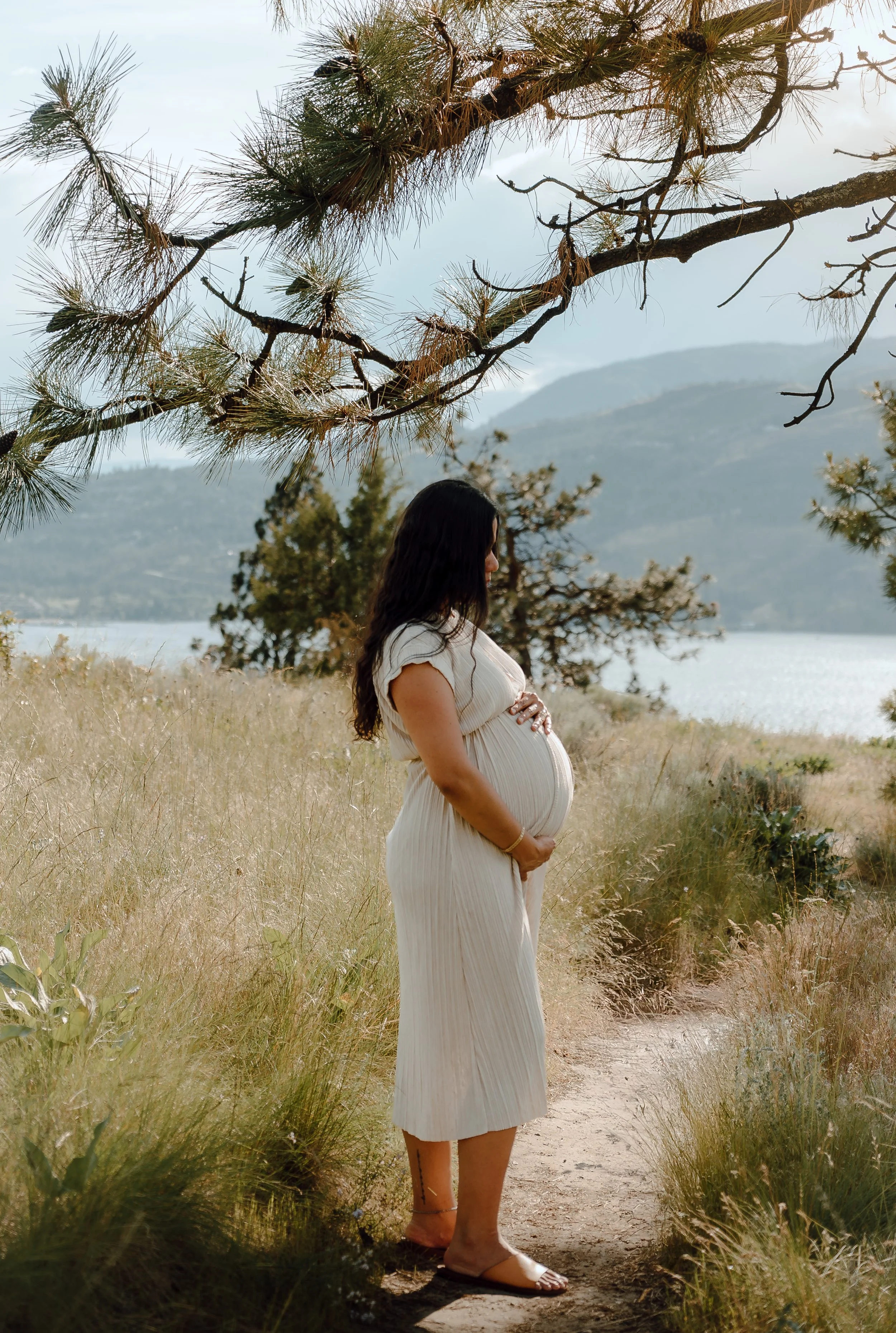 Pregnant woman standing on a trail next to a lake, surrounded by grass and trees, with mountains in the background.