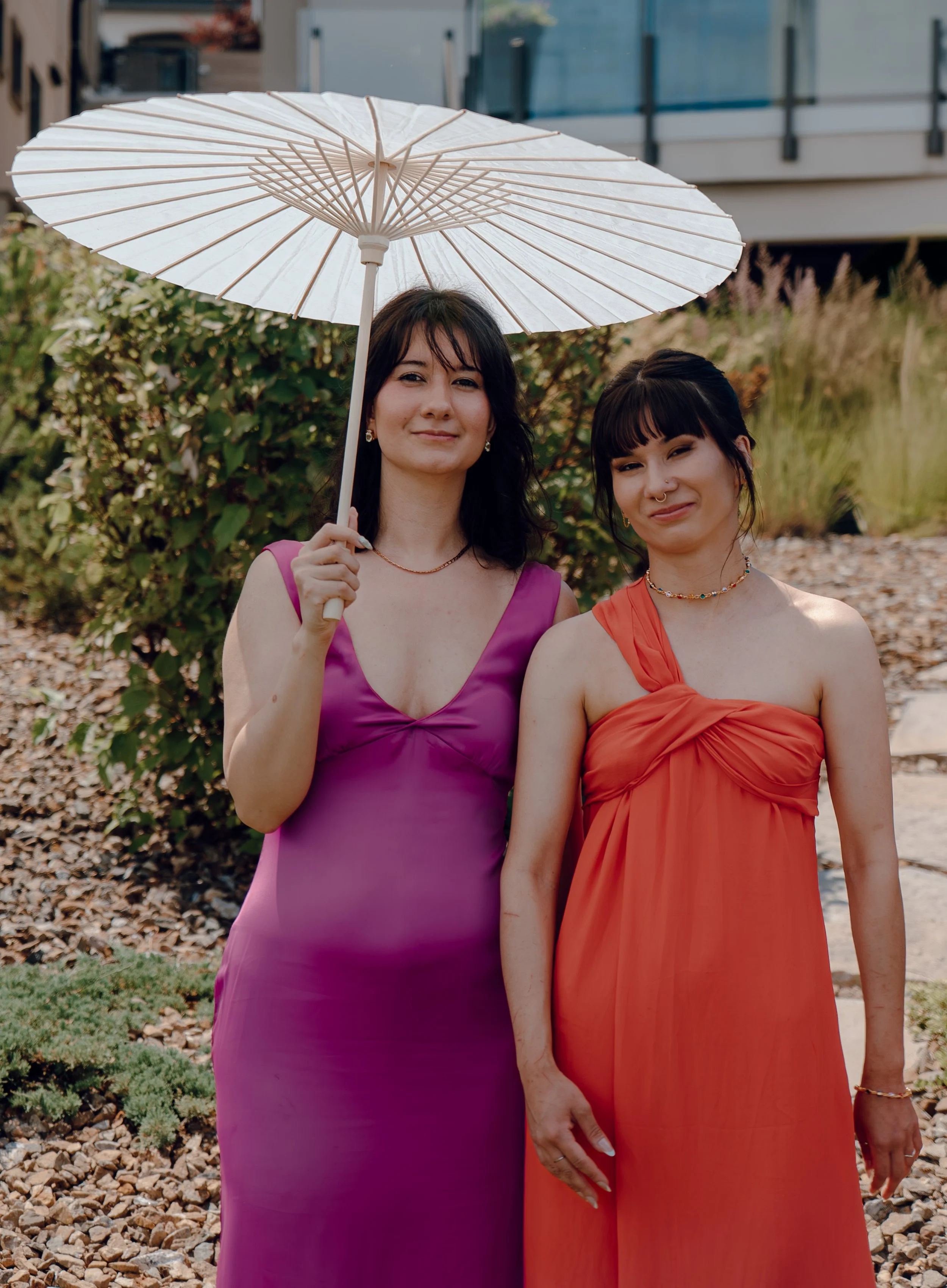 Two women standing outdoors, one holding a white parasol, both wearing colorful dresses, surrounded by greenery and rocks.