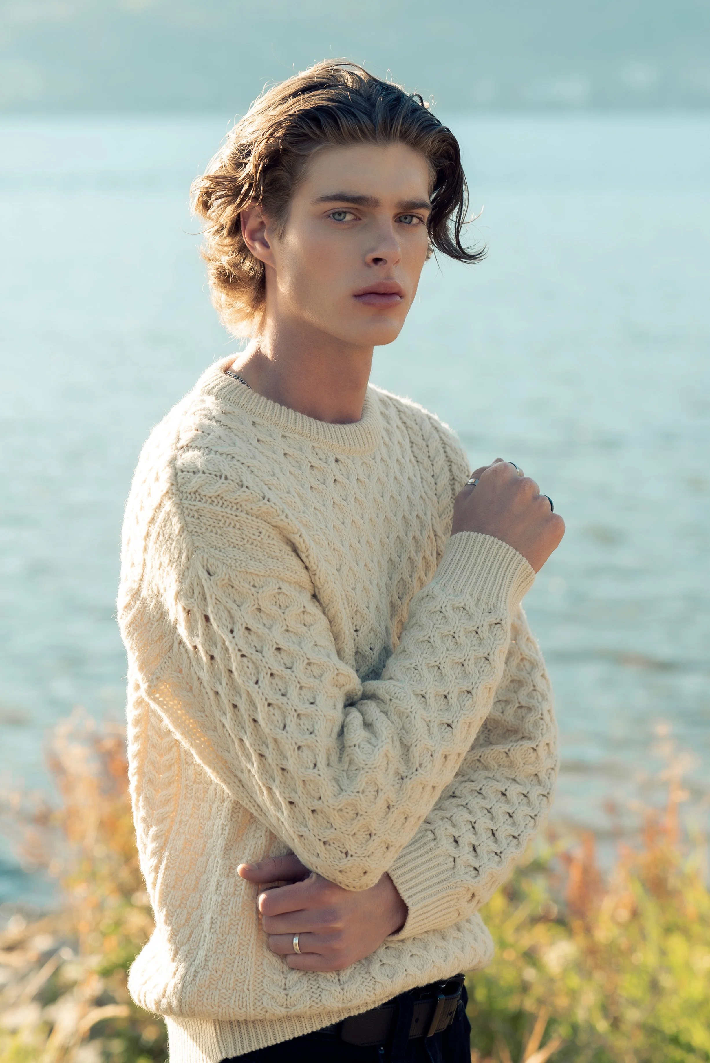 A young man with wavy hair looking at the camera stands outdoors near a body of water, wearing a cream-colored knitted sweater.