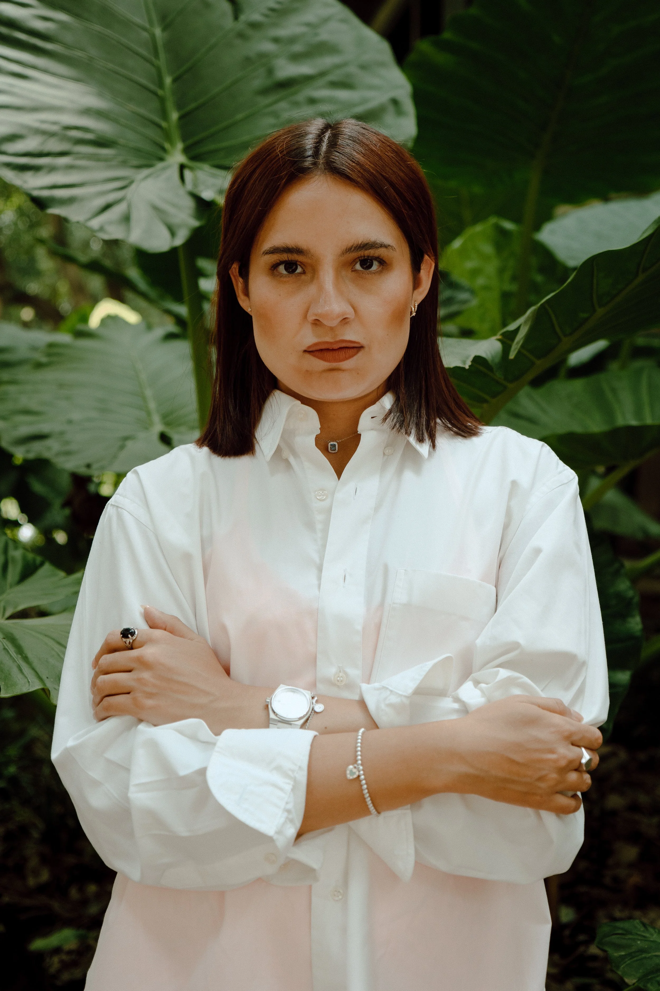 A woman with shoulder-length dark hair and light skin, wearing a white button-up shirt, standing with arms crossed in front of large green leaves.