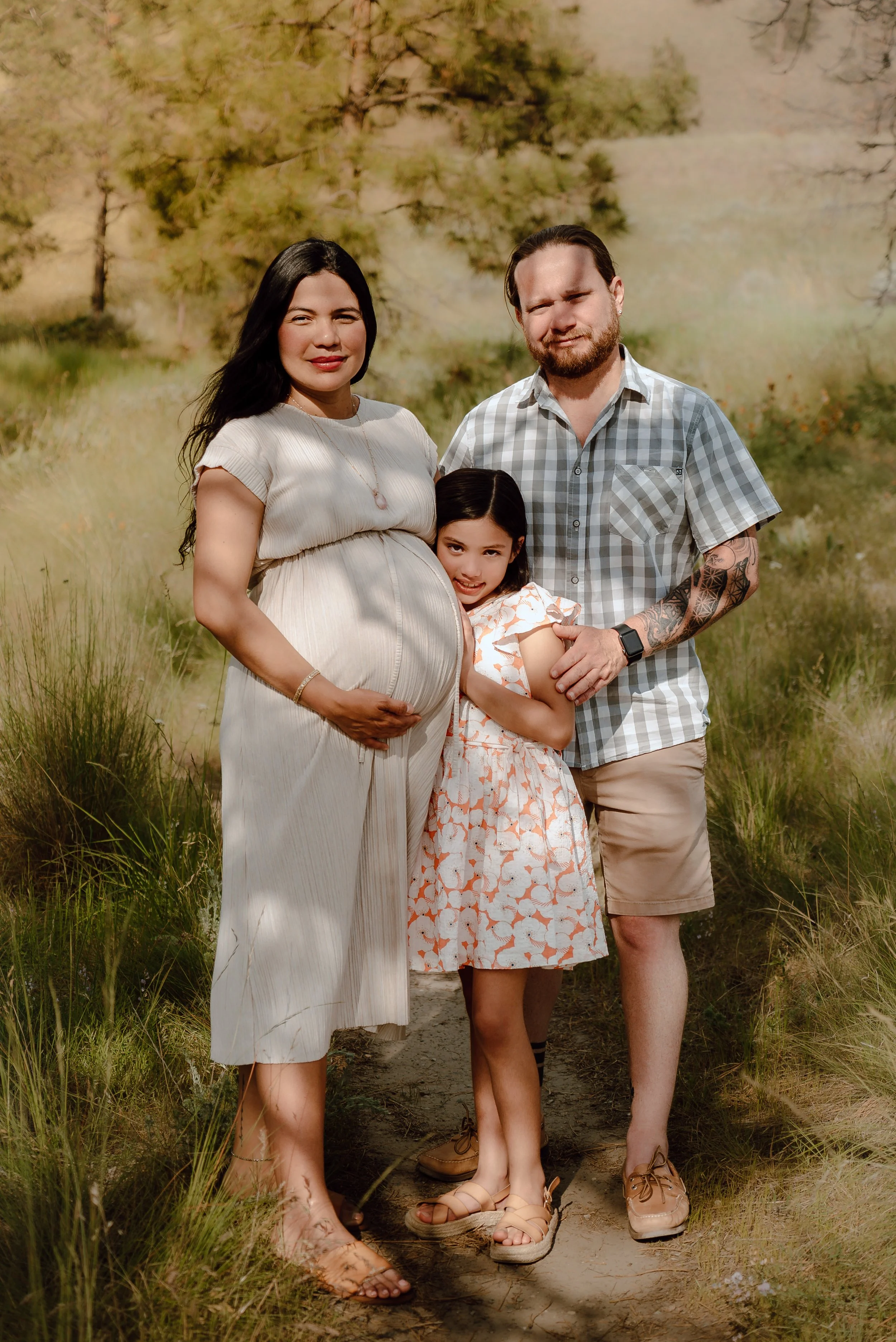 A pregnant woman, an adult man, and a young girl standing outdoors on a dirt path surrounded by grass and trees in a natural setting.