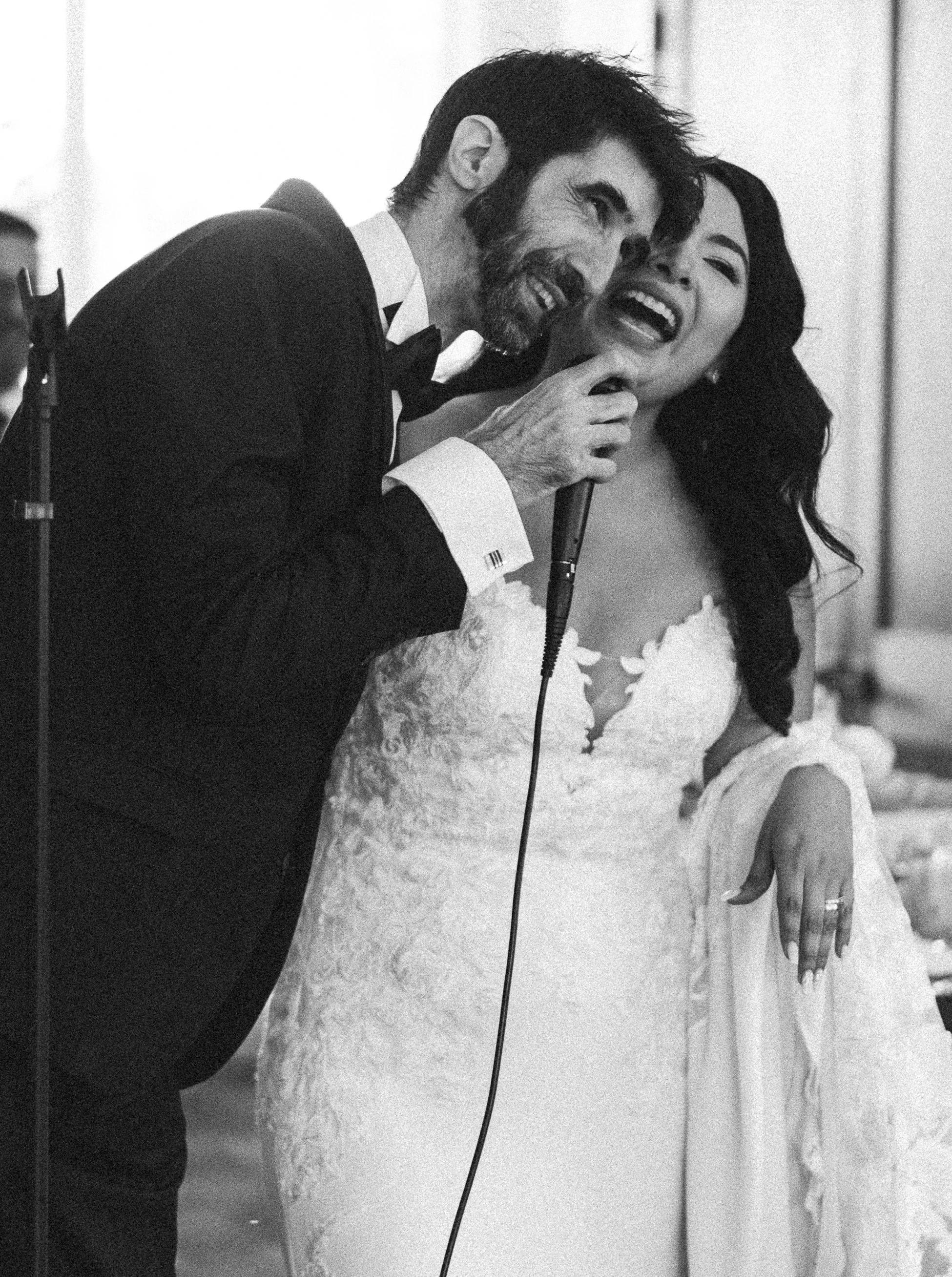A black-and-white photo of a man in a tuxedo singing into a microphone, with a woman in a wedding dress smiling and holding him.