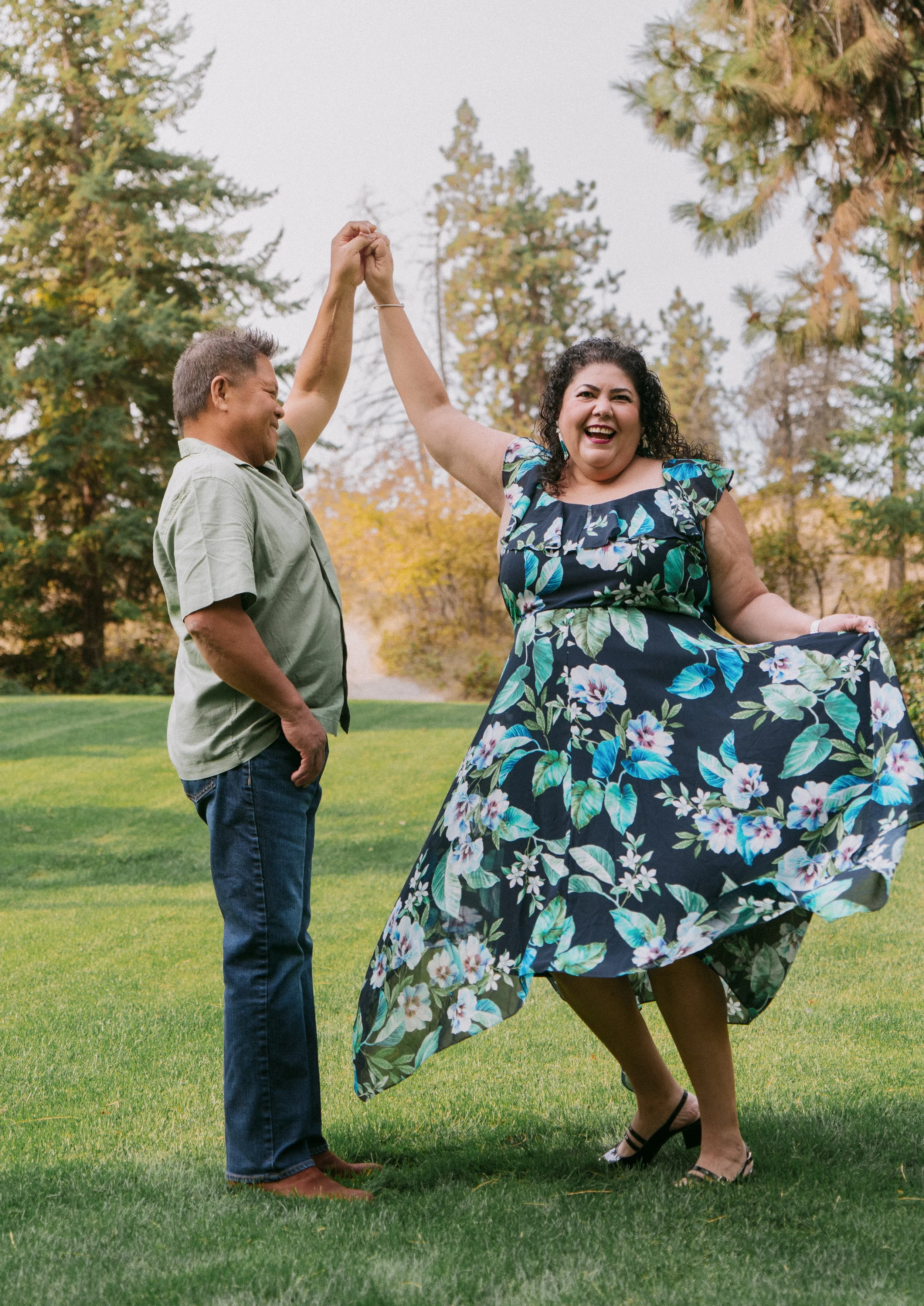 A happy woman in a floral dress being twirled by a man in a green shirt and jeans in a park with green grass and trees.