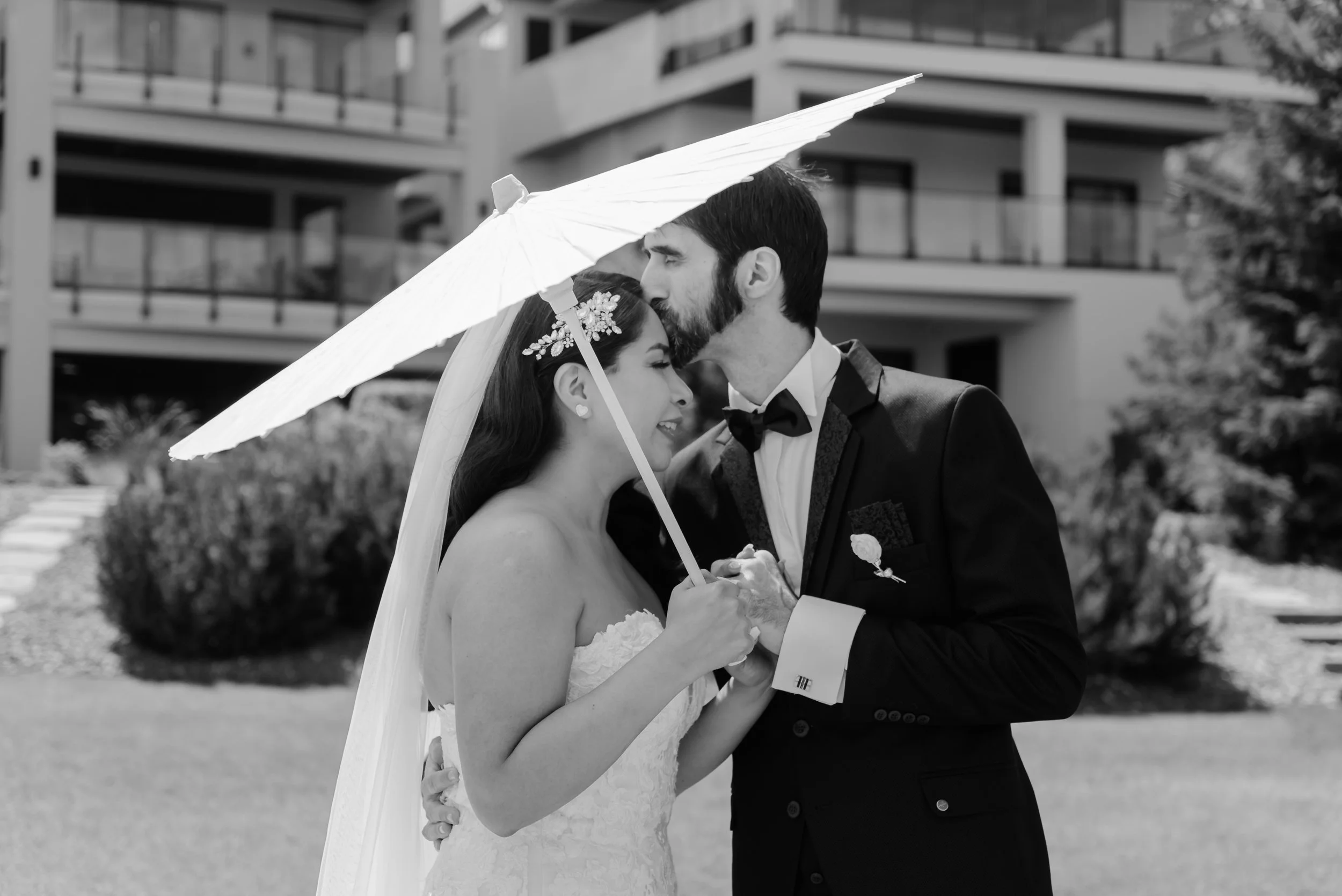 A black-and-white photo of a newlywed couple outdoors, with the groom kissing the bride on her forehead while holding a parasol over them. The bride is wearing a strapless wedding gown and the groom is dressed in a tuxedo with a bow tie.