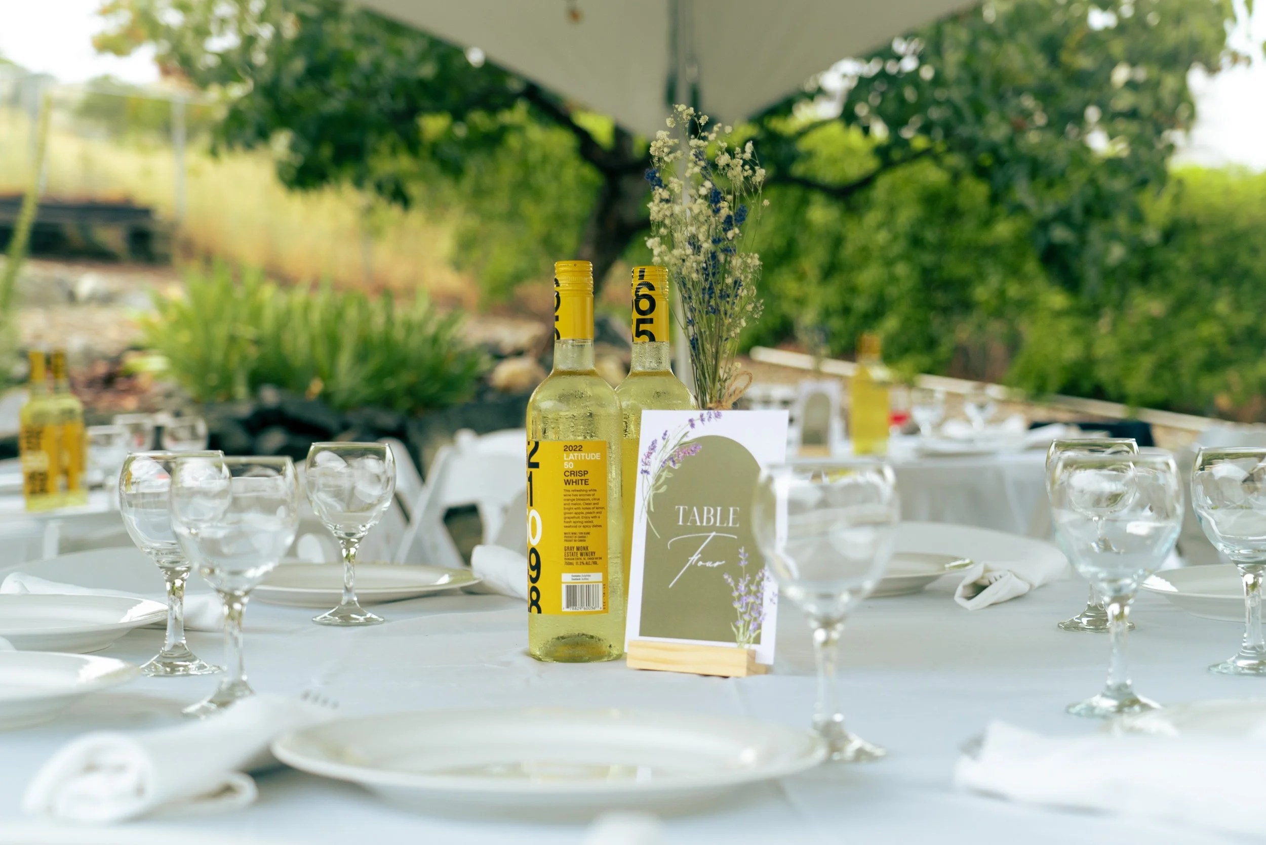 Table set for an outdoor event with wine bottles, glasses, a table card, and floral decorations.