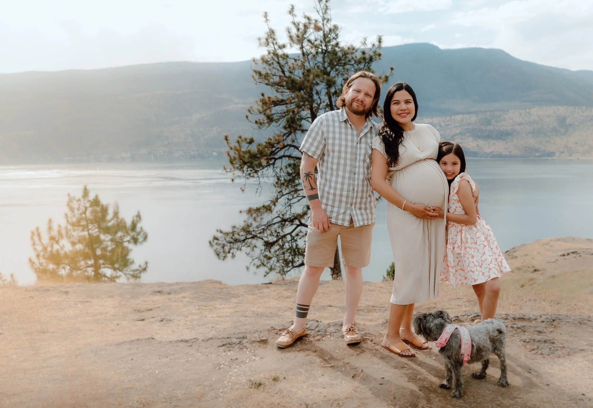 A family of three plus a dog standing outdoors by a lake with mountains in the background. The pregnant woman is in the center, wearing a light dress, with her hand on her belly. The man is to her left, wearing a checkered shirt and kh shorts, with a