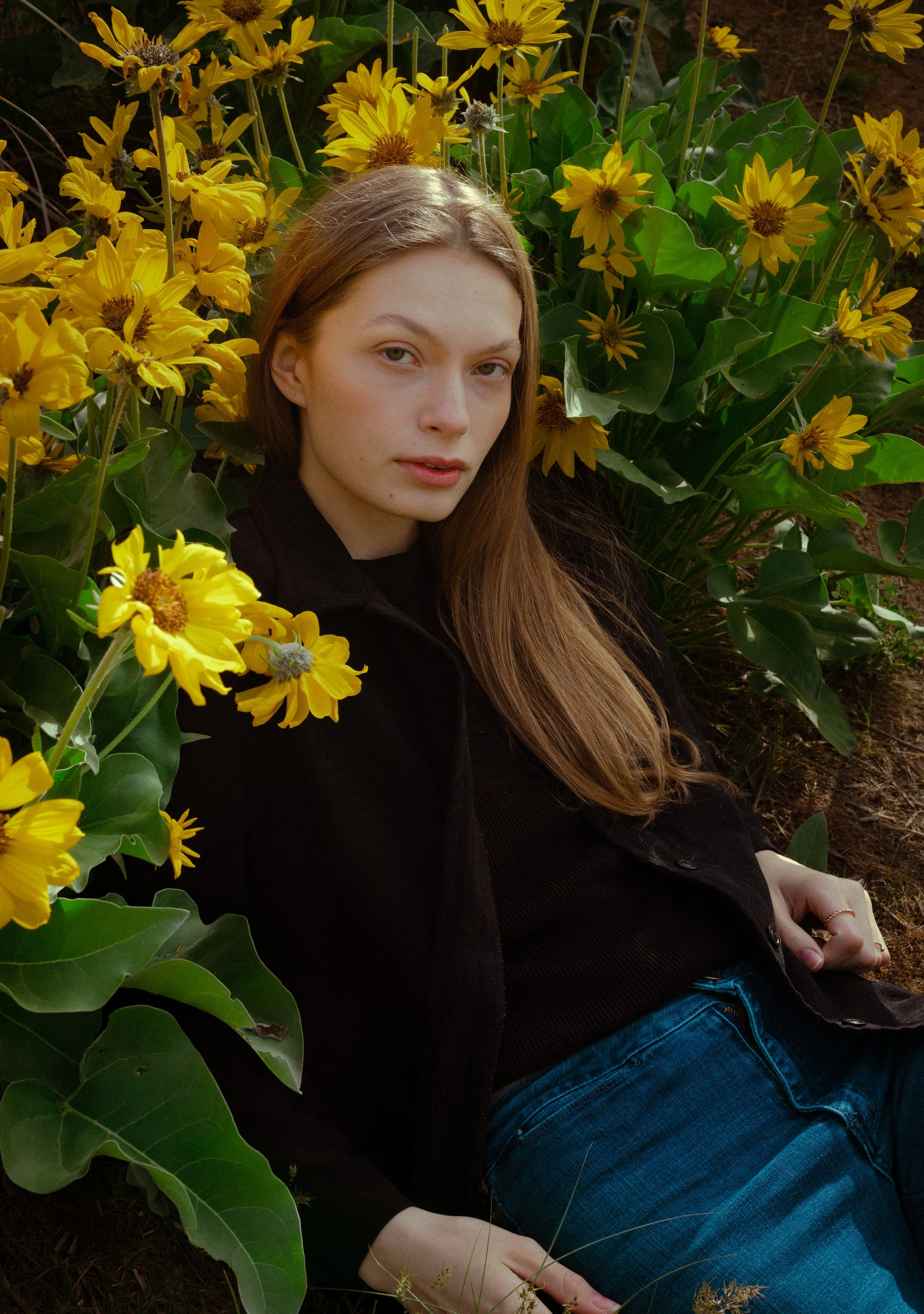 A young woman with long, reddish-brown hair, wearing a black top and dark jacket, sitting among yellow flowers and green leaves.