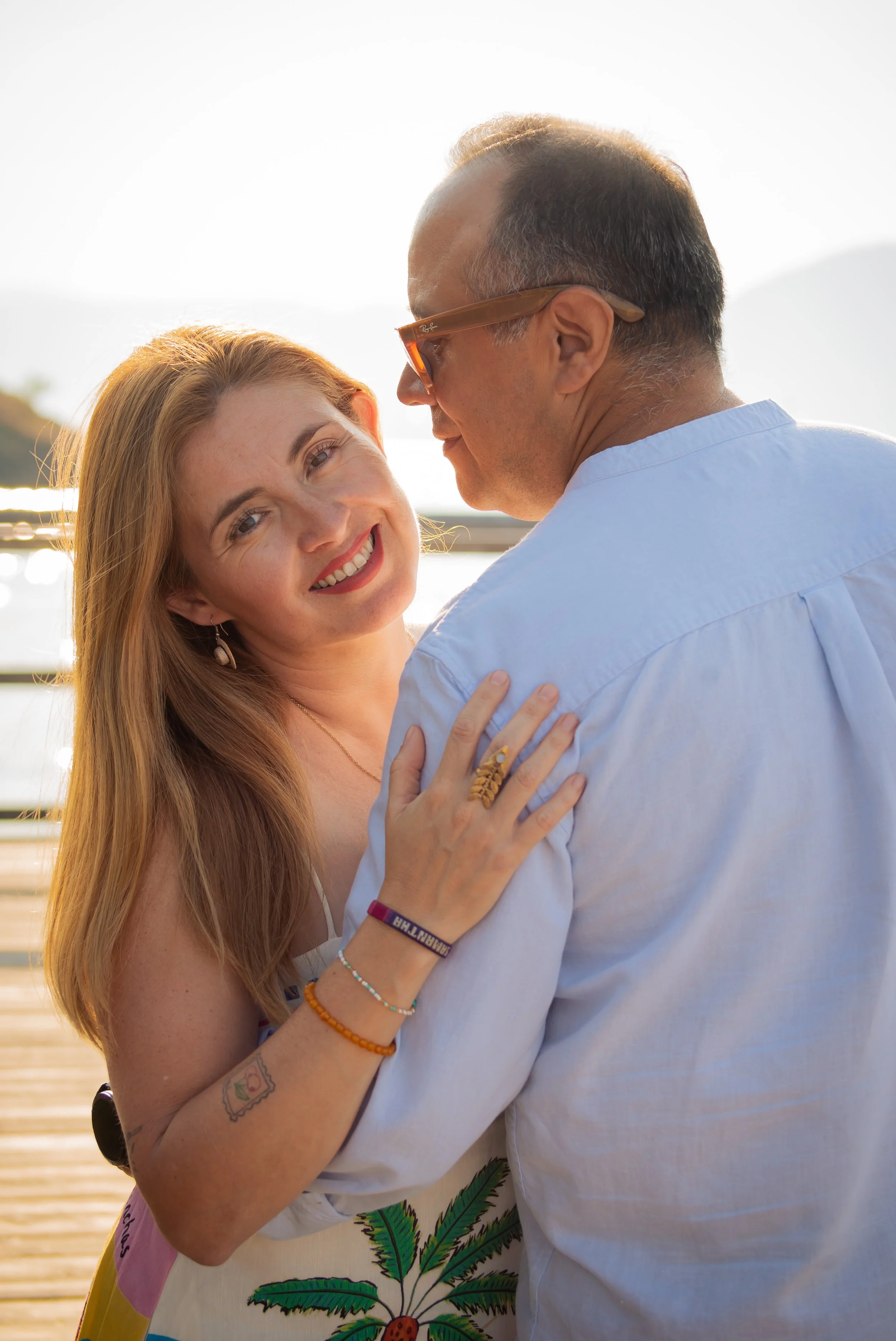 A smiling woman with long red hair and a man with sunglasses sharing an embrace outdoors, with sunlight in the background.