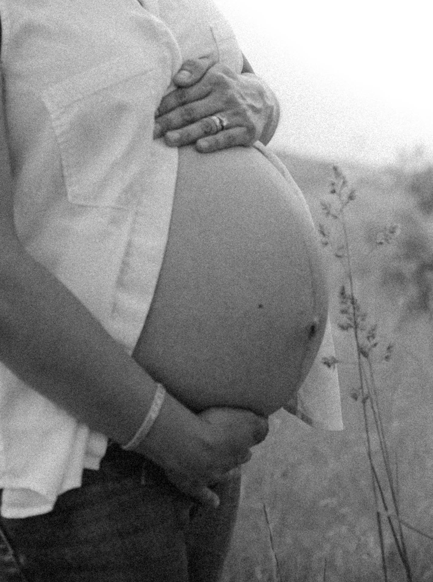 Black and white photo of a pregnant woman holding her belly with both hands, wearing a light-colored top and pants.