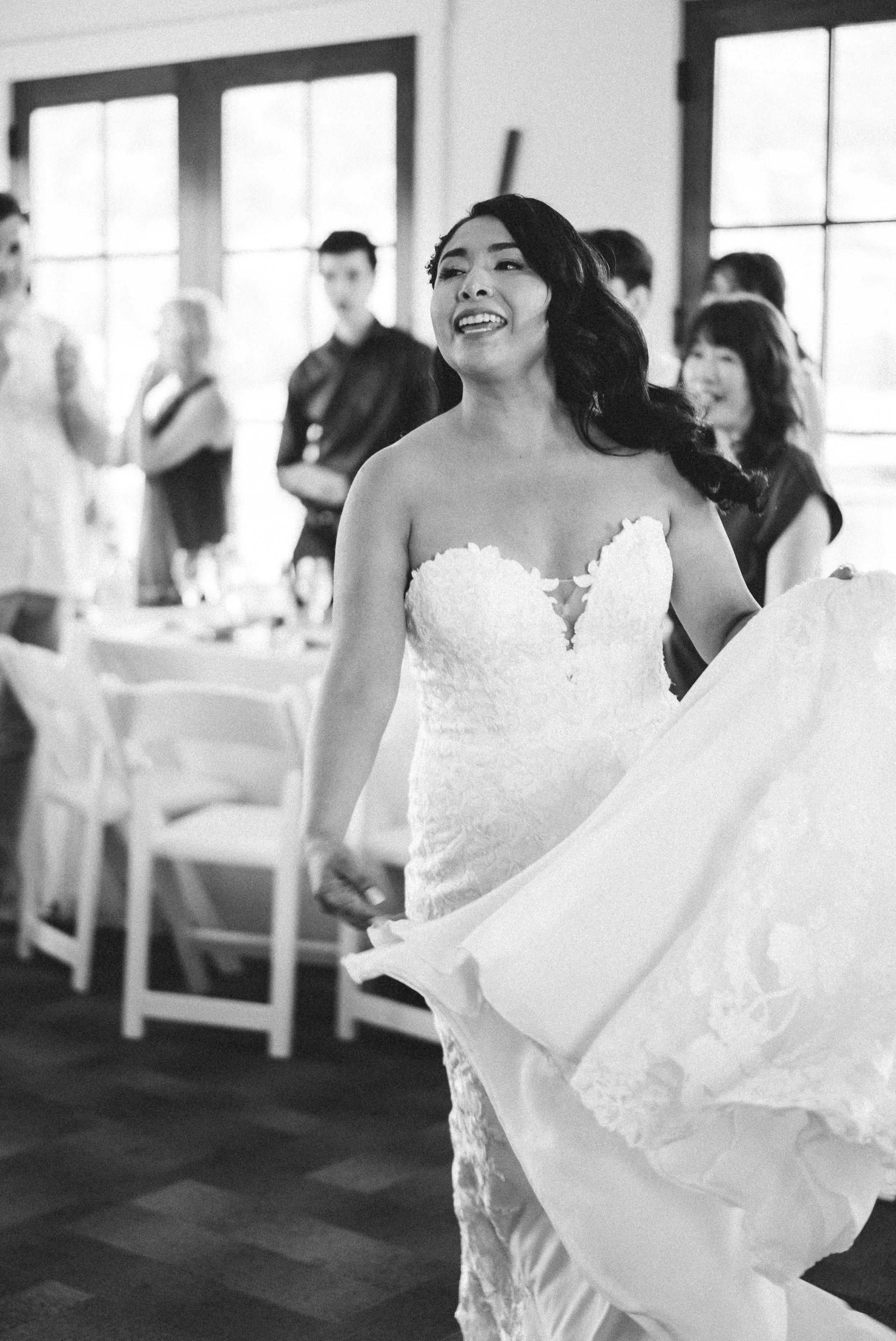 A joyful woman in a wedding dress, holding the hem of her gown, at a wedding reception with guests in the background.