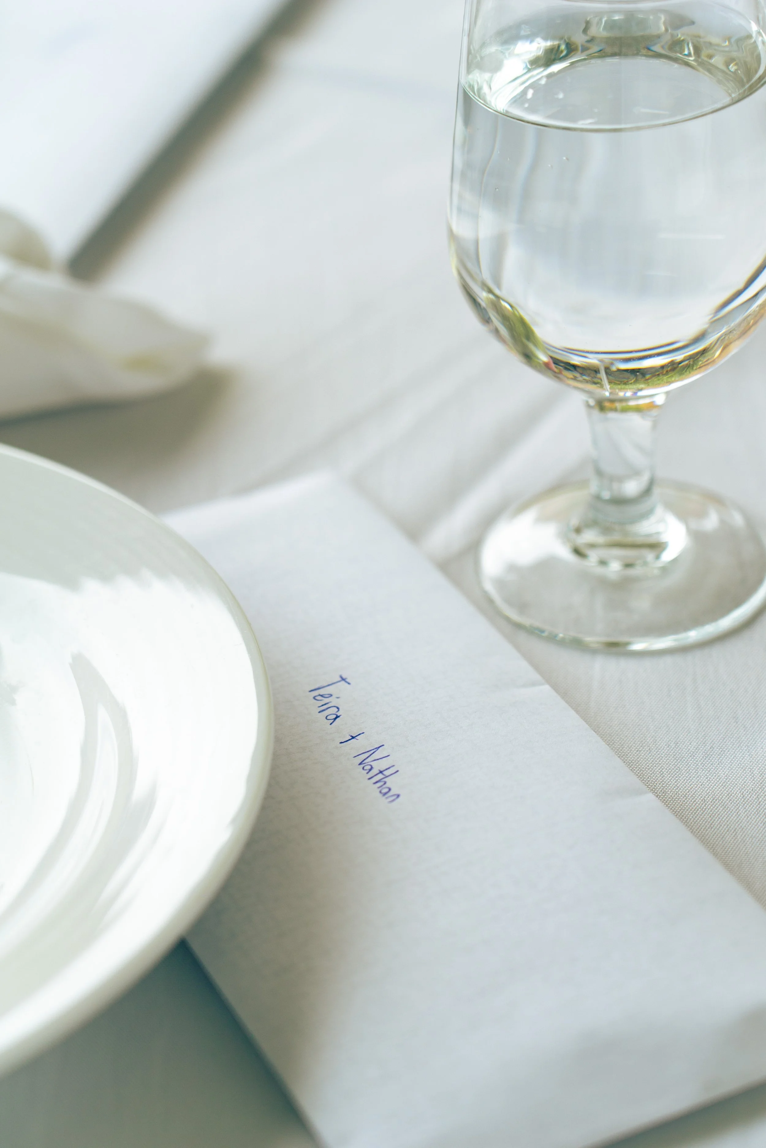 A place card with the names 'tina + nathan' written on it, set on a table next to a glass of water and a white plate.