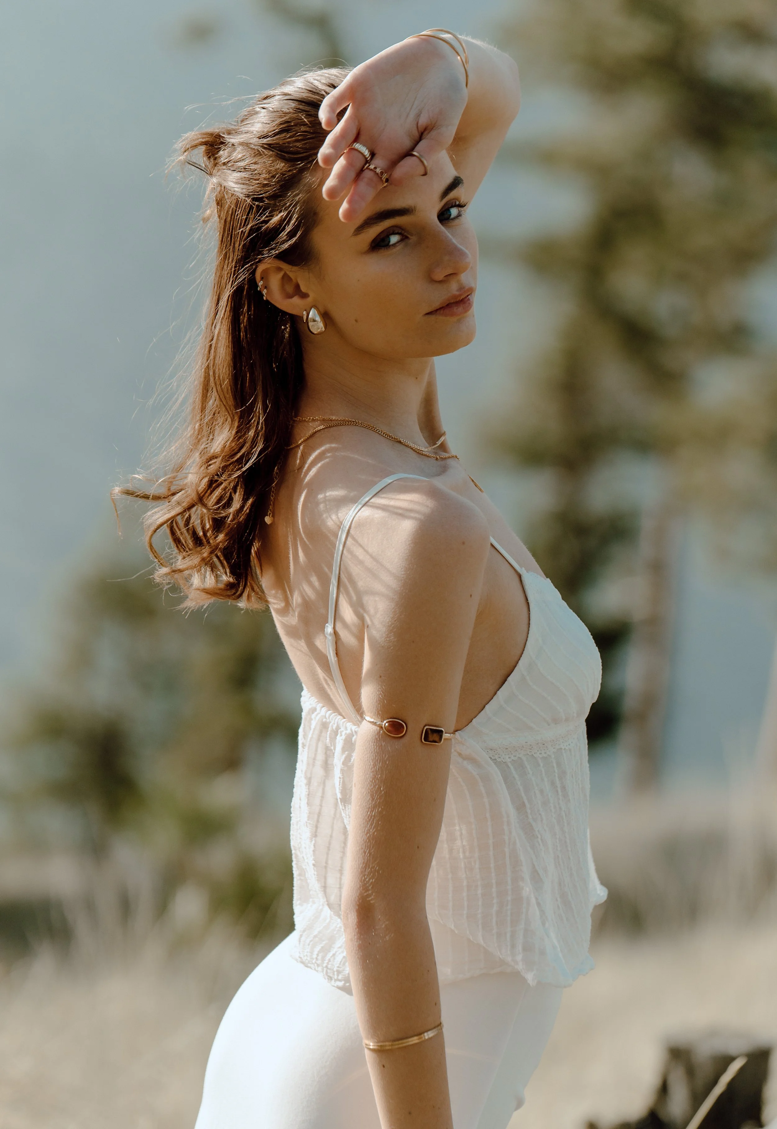 A young woman with wavy brown hair and jewelry, wearing a white sleeveless top, standing outdoors with blurred trees and water in the background.